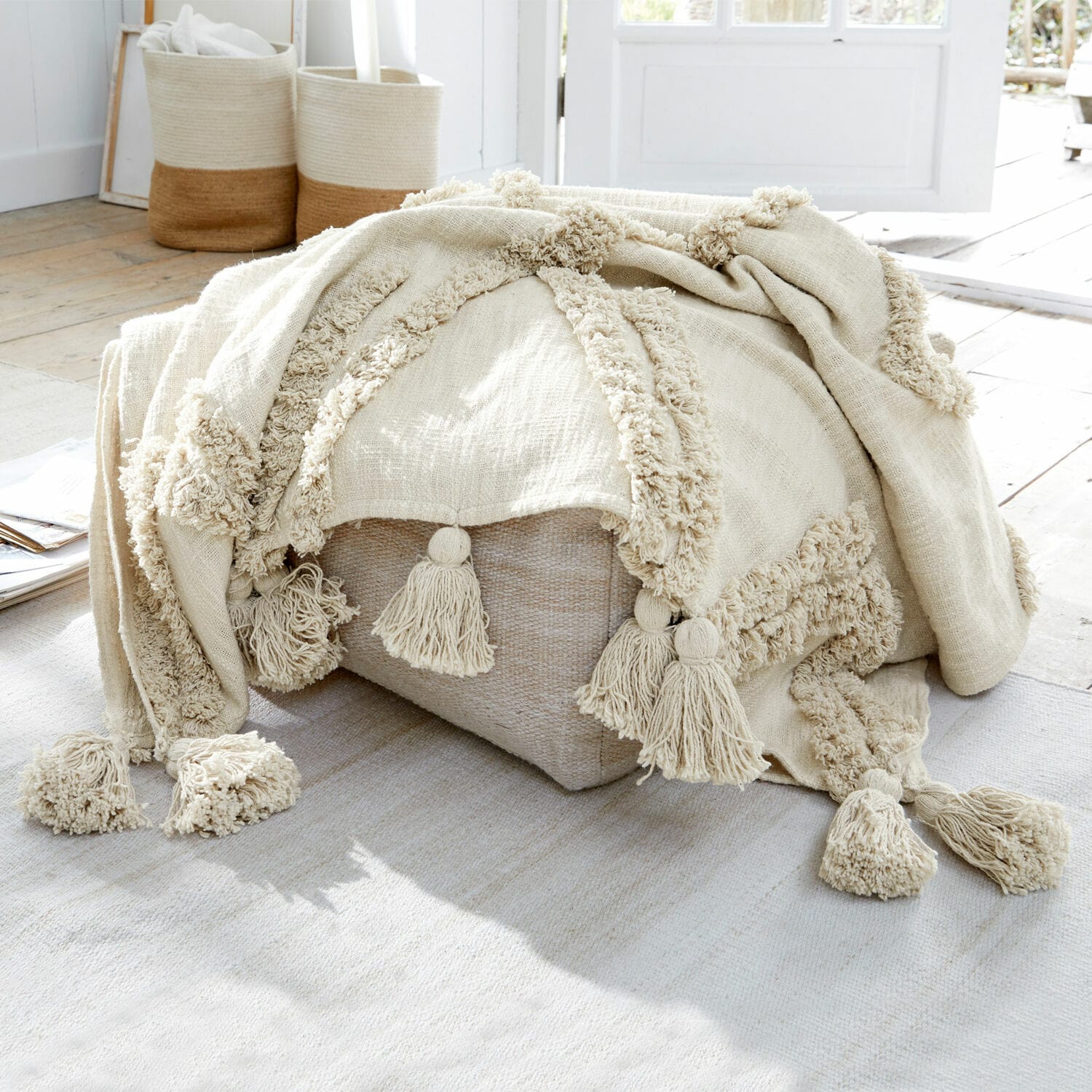 Plaid Linares Plaid beige en coton avec pompons sur un pouf clair dans un intérieur lumineux naturel