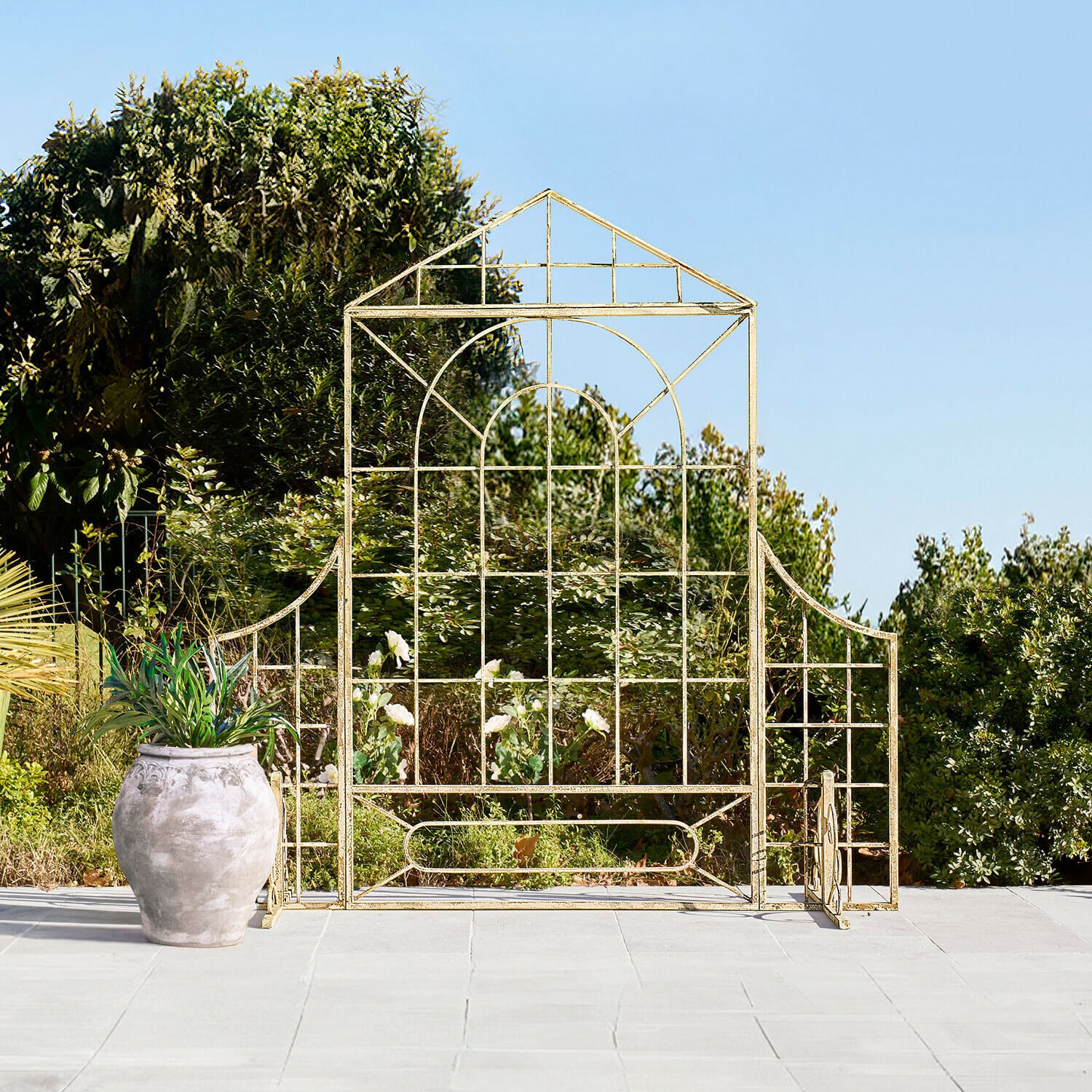 Paravent Hallowby Treillis de jardin en métal doré en forme de maison sur terrasse avec pot et végétation en arrière-plan