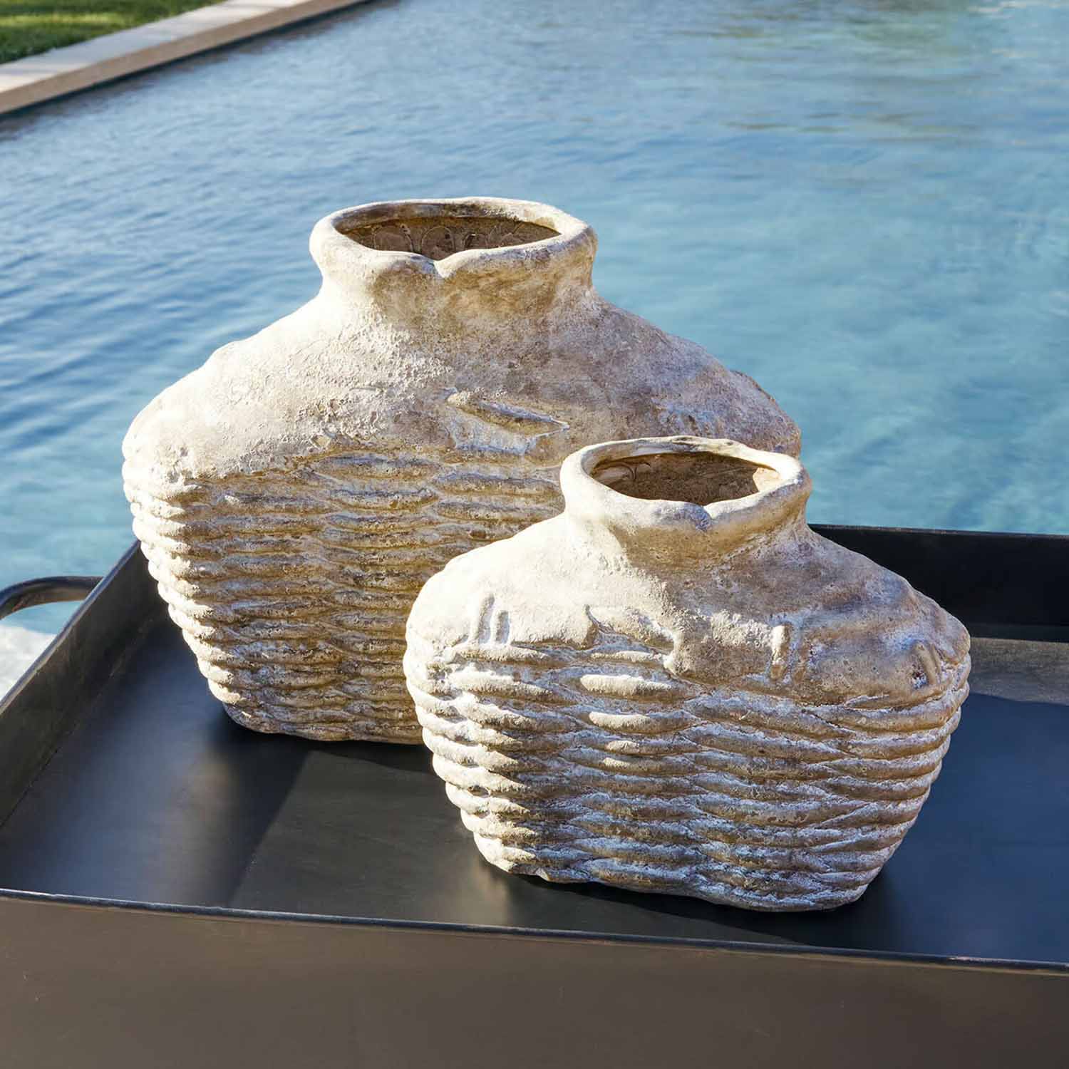 Deux vases rustiques en pierre beige posés sur un plateau noir près d'une piscine