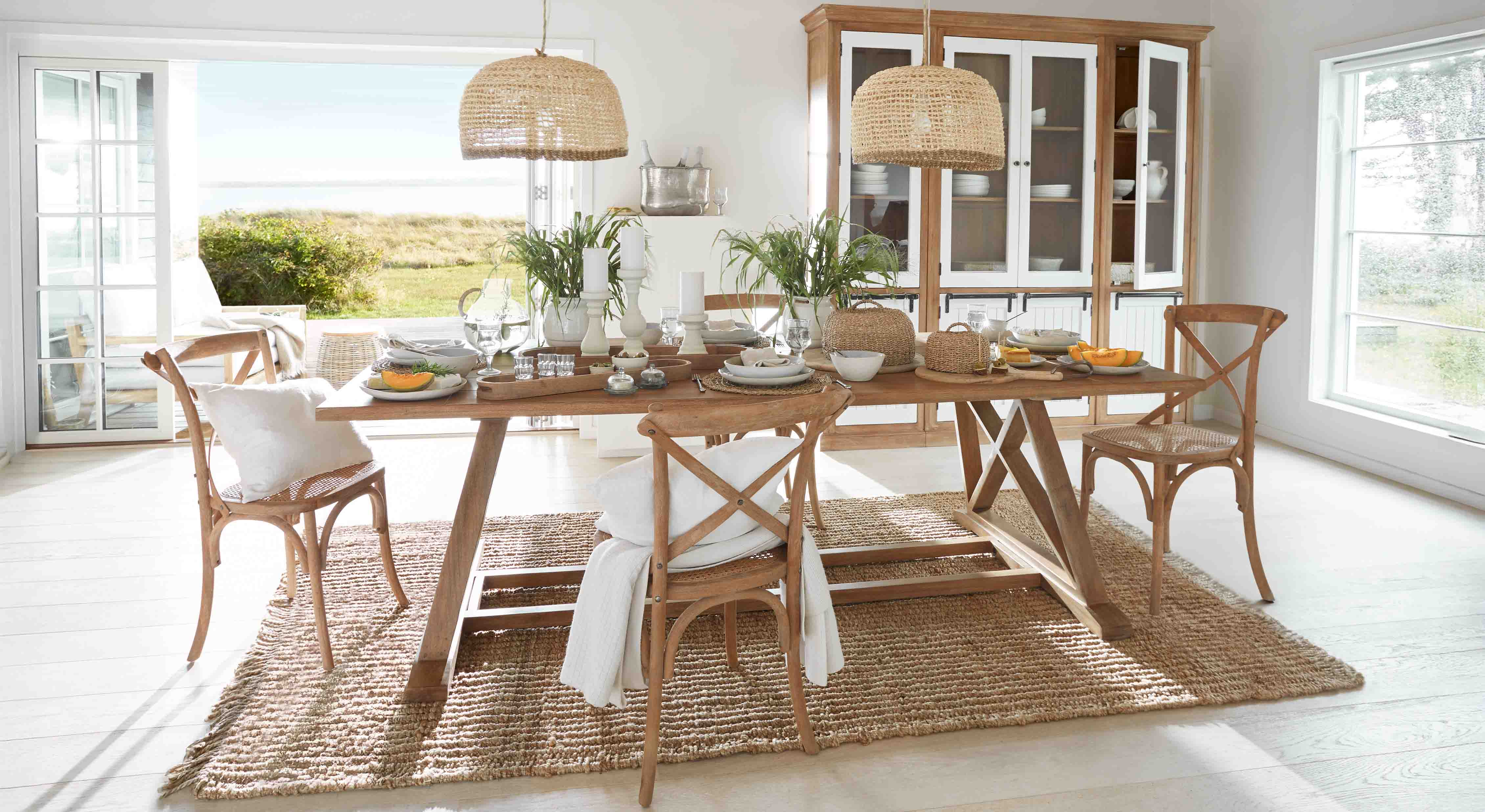 Eetkamer met houten tafel en rotan stoelen op natuurlijke vloerkleed, planten en rieten hanglampen