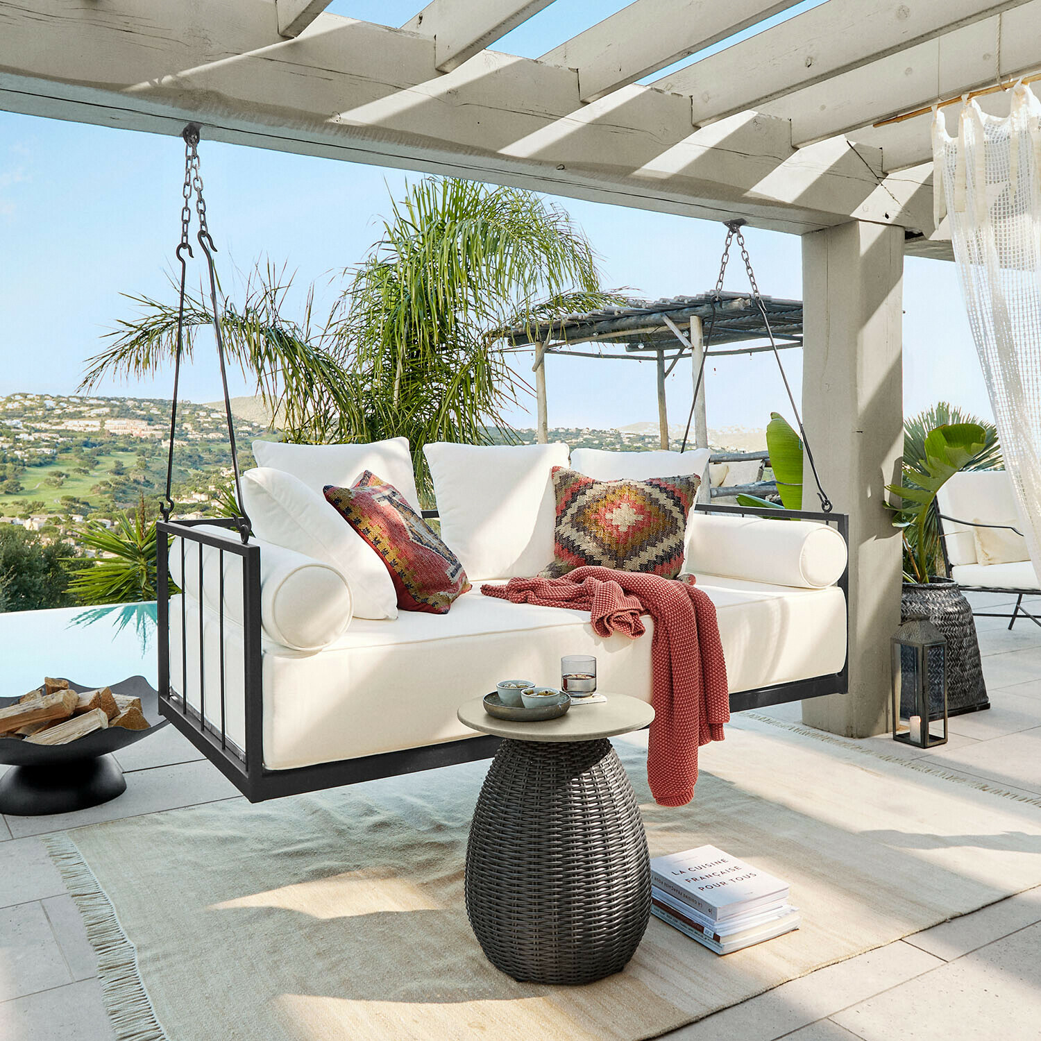 Banquette suspendue Bellecôtes Canapé suspendu blanc avec cadre en métal et coussins colorés sur terrasse avec vue verdoyante