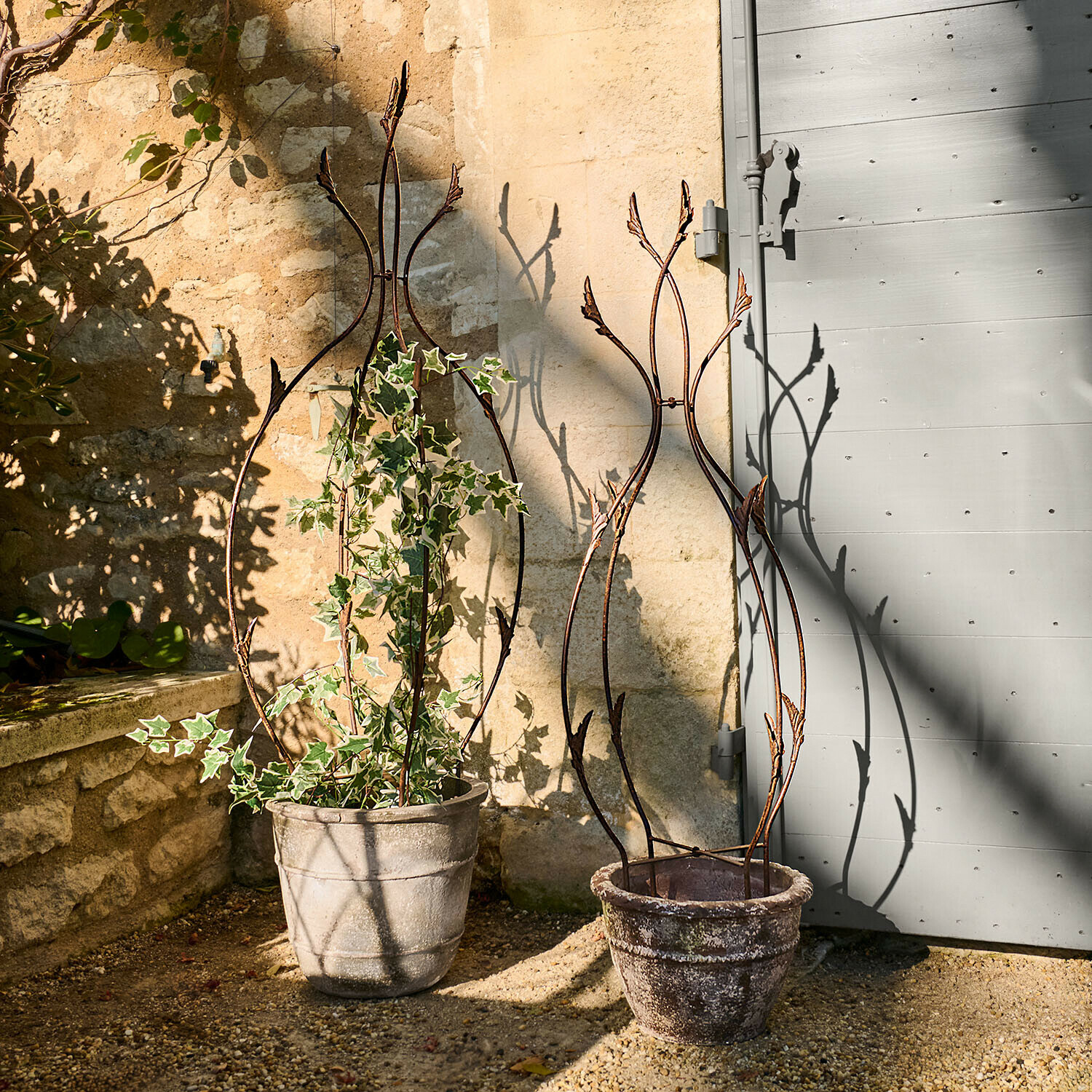 Twee decoratieve metalen plantensteunen in stenen potten op een zonnig terras