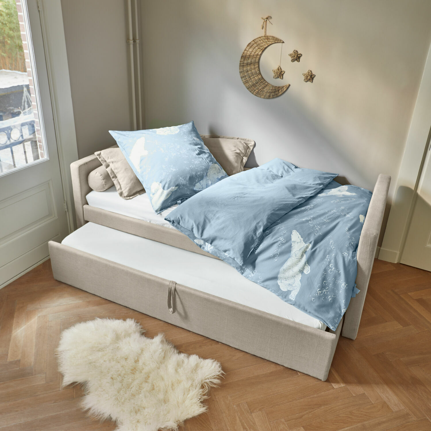 Bed Thistlewood Beige bed met uitschuifmatras en blauw beddengoed met ijsbeerprint in lichte kamer.