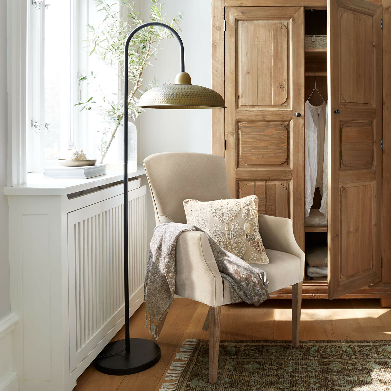 Beige fauteuil met houten poten, sierkussen en deken, naast metalen vloerlamp met messing kap in lichte kamer