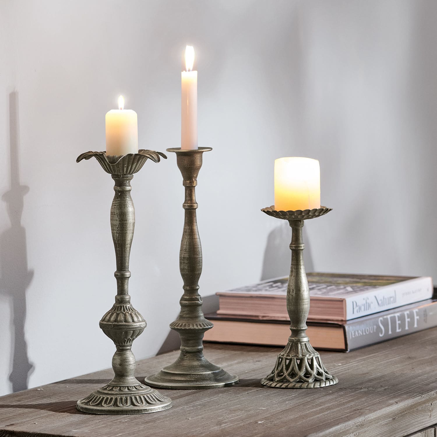 Lot de 3 bougeoirs Chandra Trois bougeoirs en métal couleur argent vieilli avec bougies allumées sur une table en bois