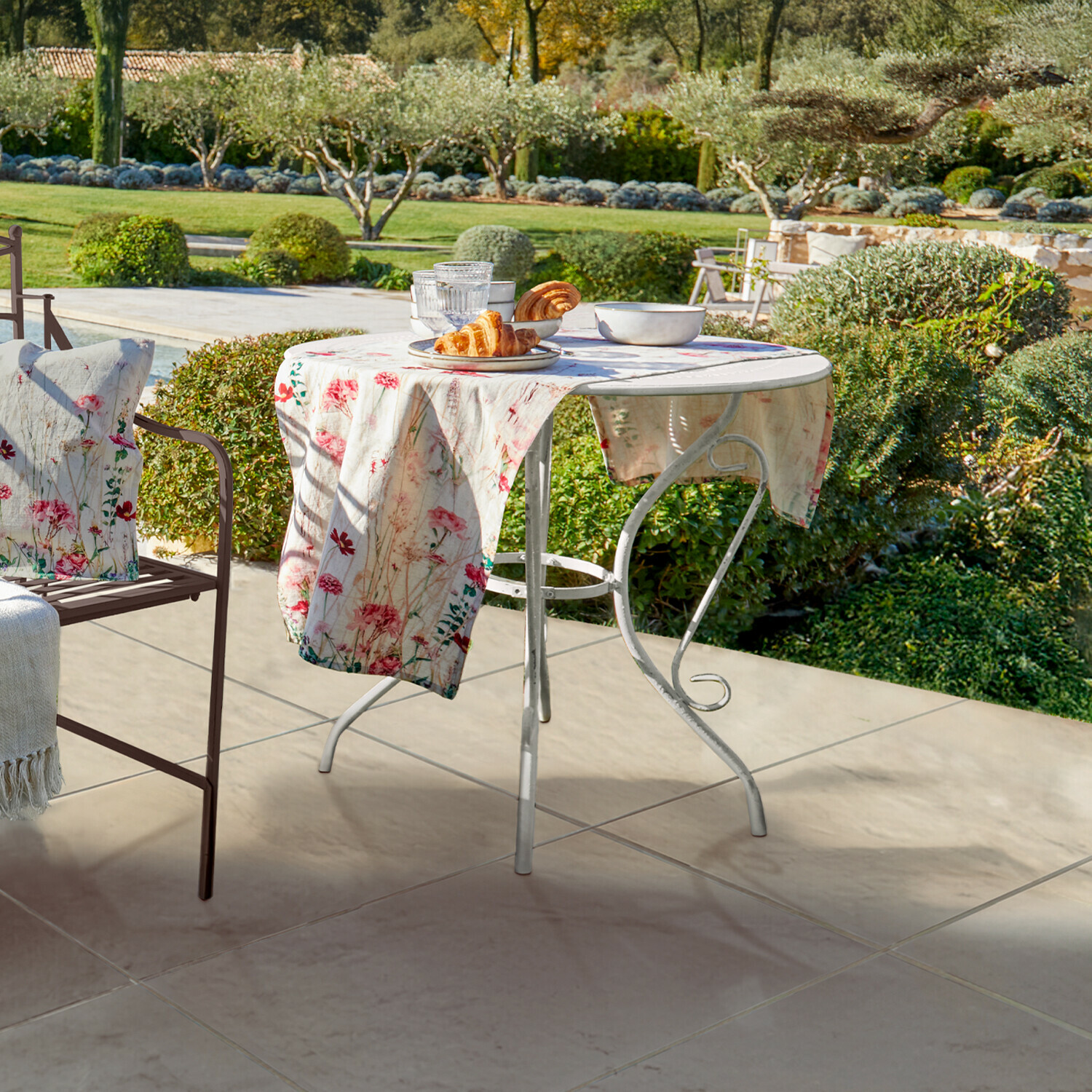 Table Aube Table de jardin ronde en métal blanc avec nappe fleurie et viennoiseries sur terrasse verdoyante