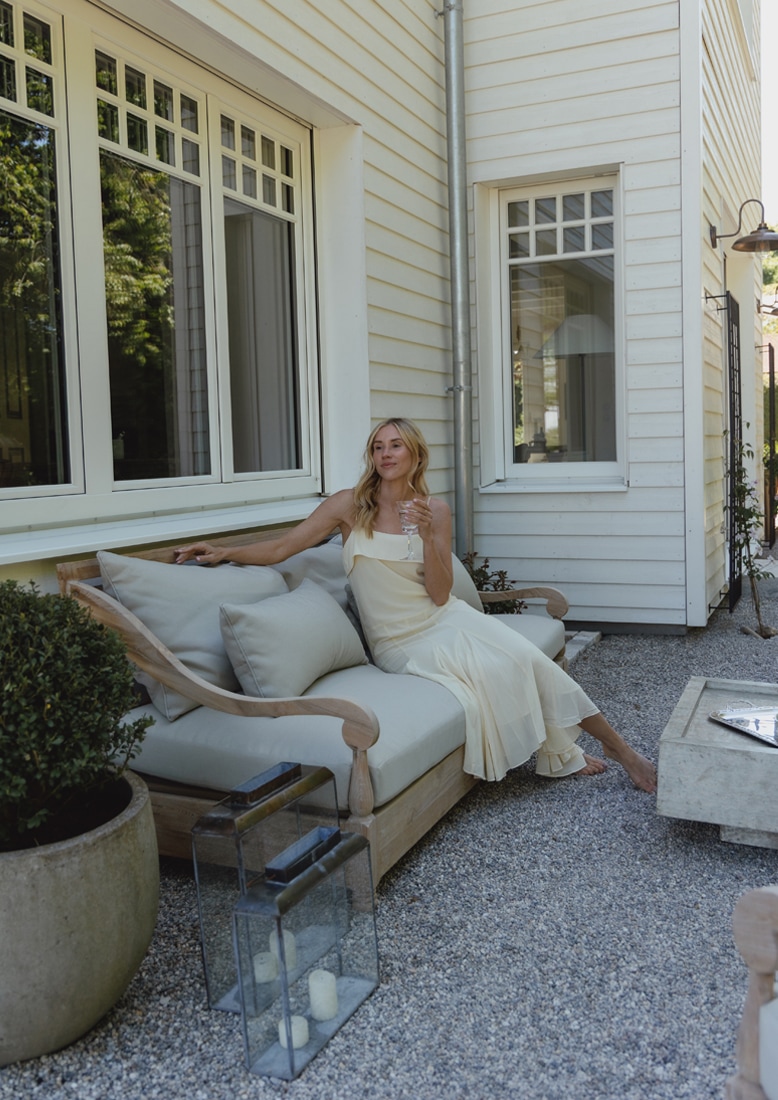 Houten loungesofa met lichtgrijze kussens op grindterras, vrouw in crèmekleurige jurk