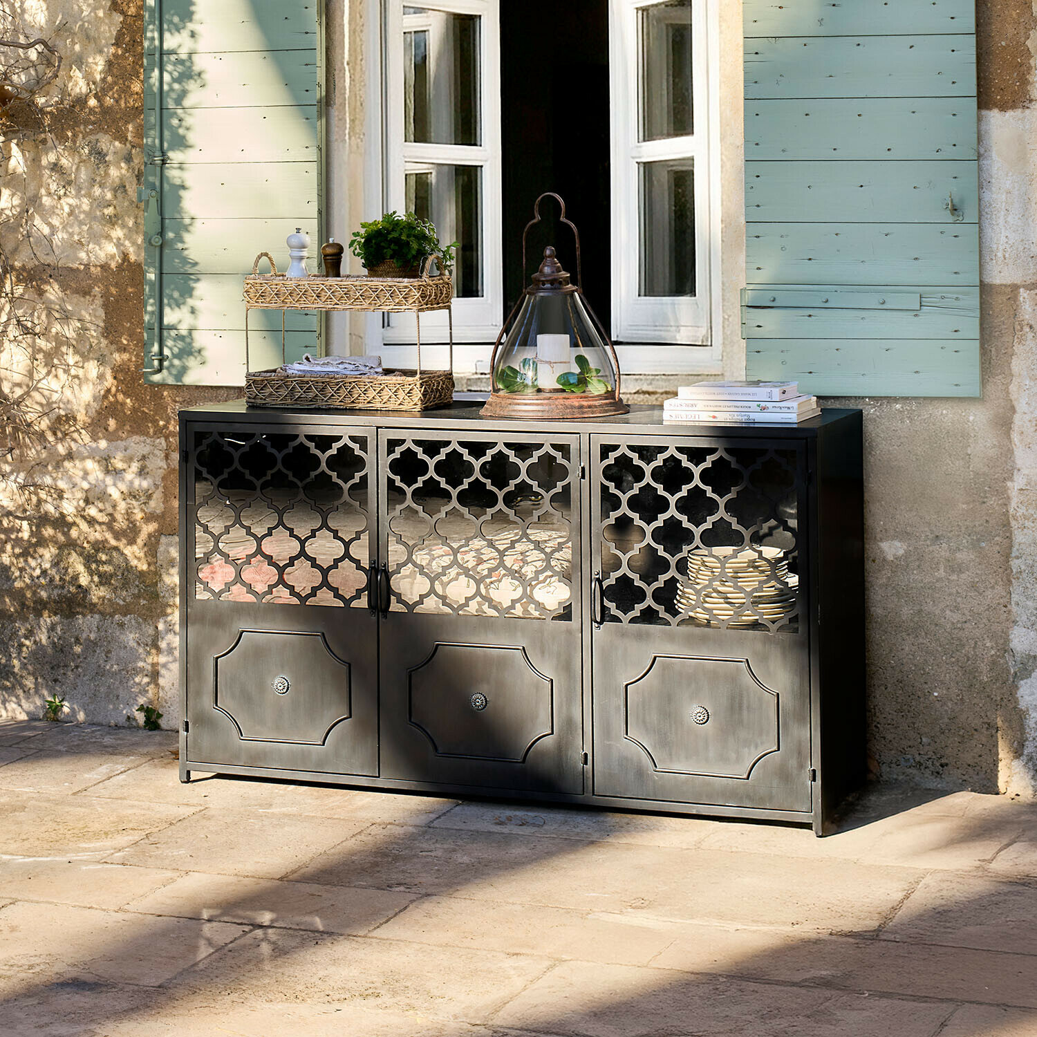 Buffet bas Astralon Buffet noir en métal avec portes ajourées sur une terrasse devant une fenêtre aux volets verts