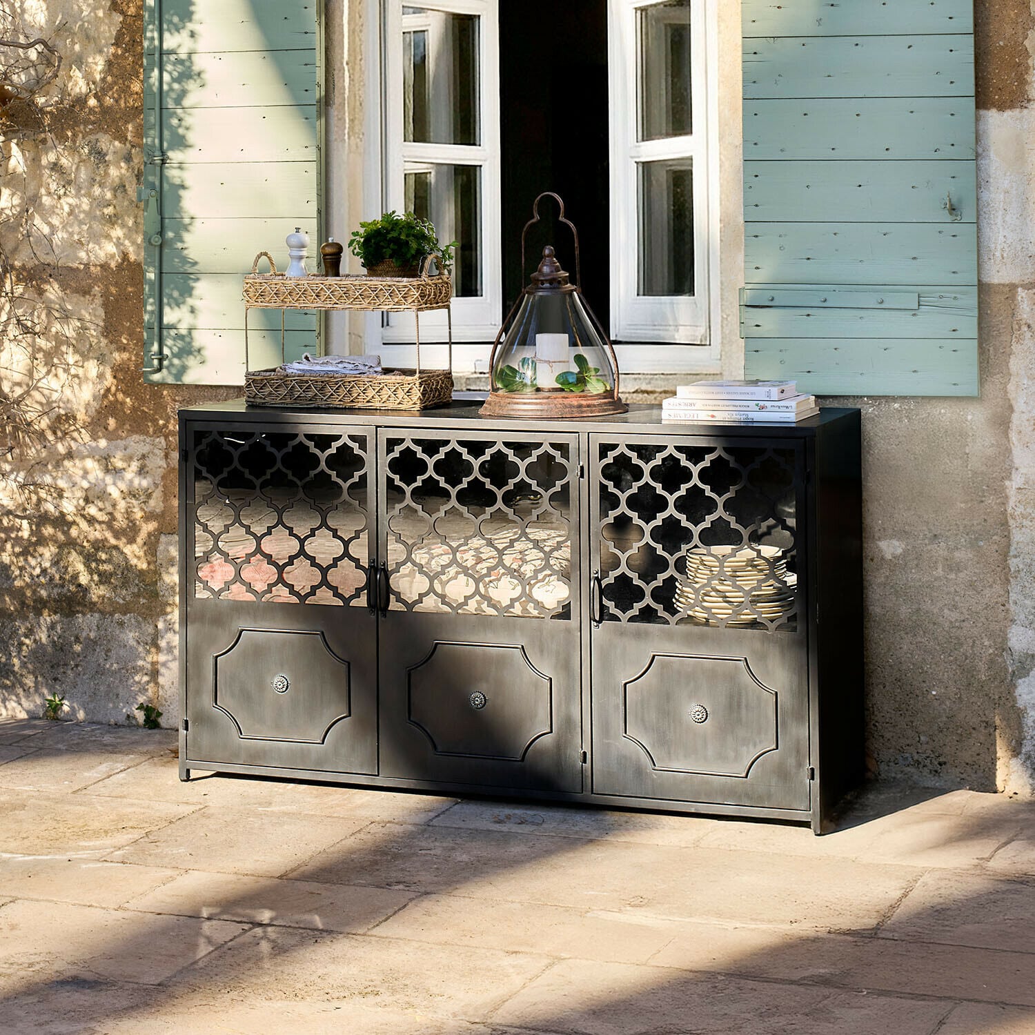 Dressoir Astralon Zwart metalen dressoir met geperforeerde deuren op terras voor raam met groene luiken