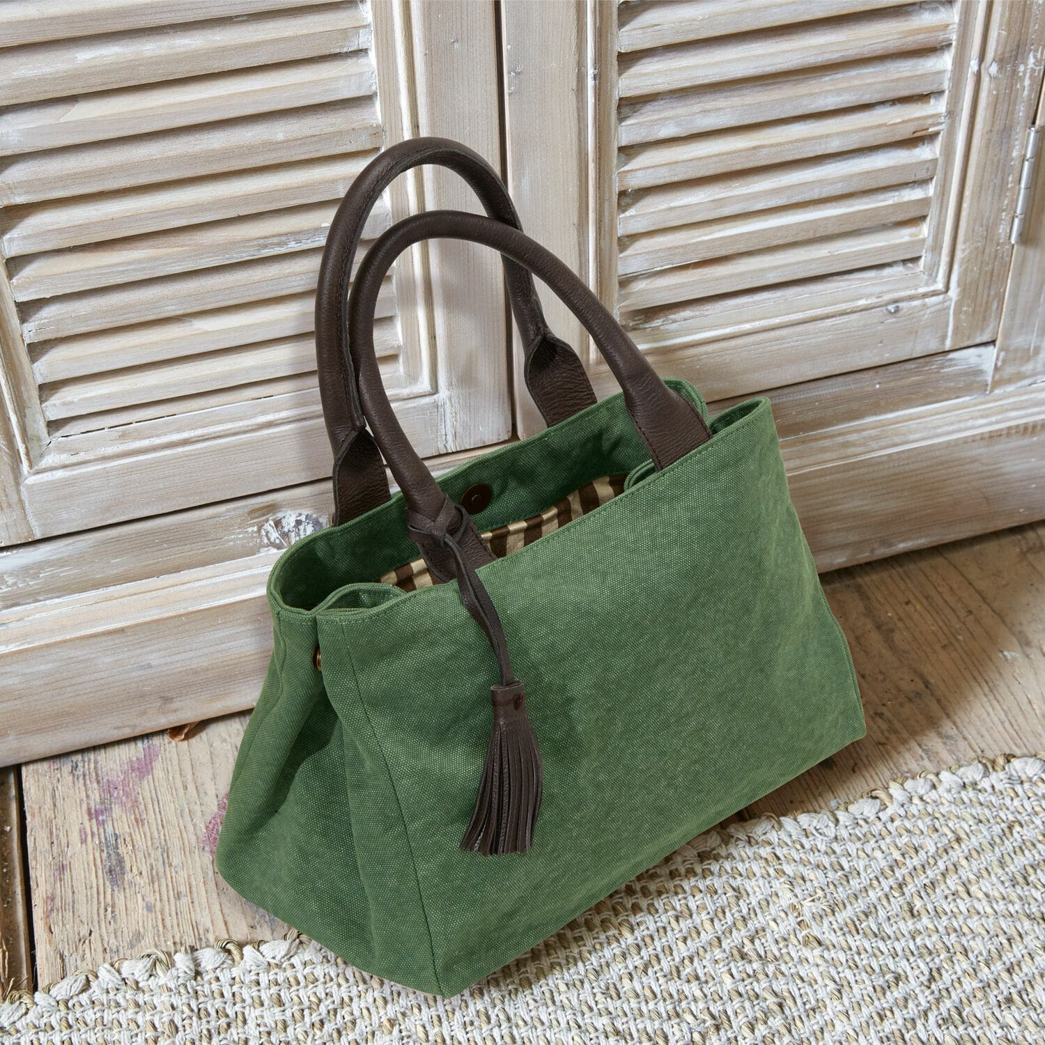 Sac Hoya Sac vert en tissu avec poignées en cuir marron et pompon posé sur un sol en bois clair