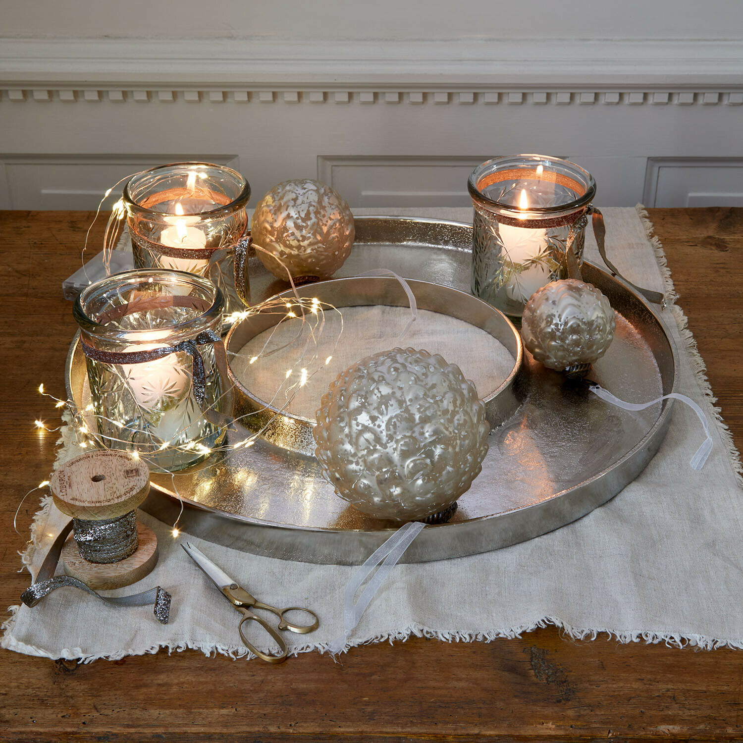 Kaarsenplateau Velarosas Zilverkleurige metalen schaal met decoratieve kaarsen en glazen ballen op houten tafel met LED-lichtjes