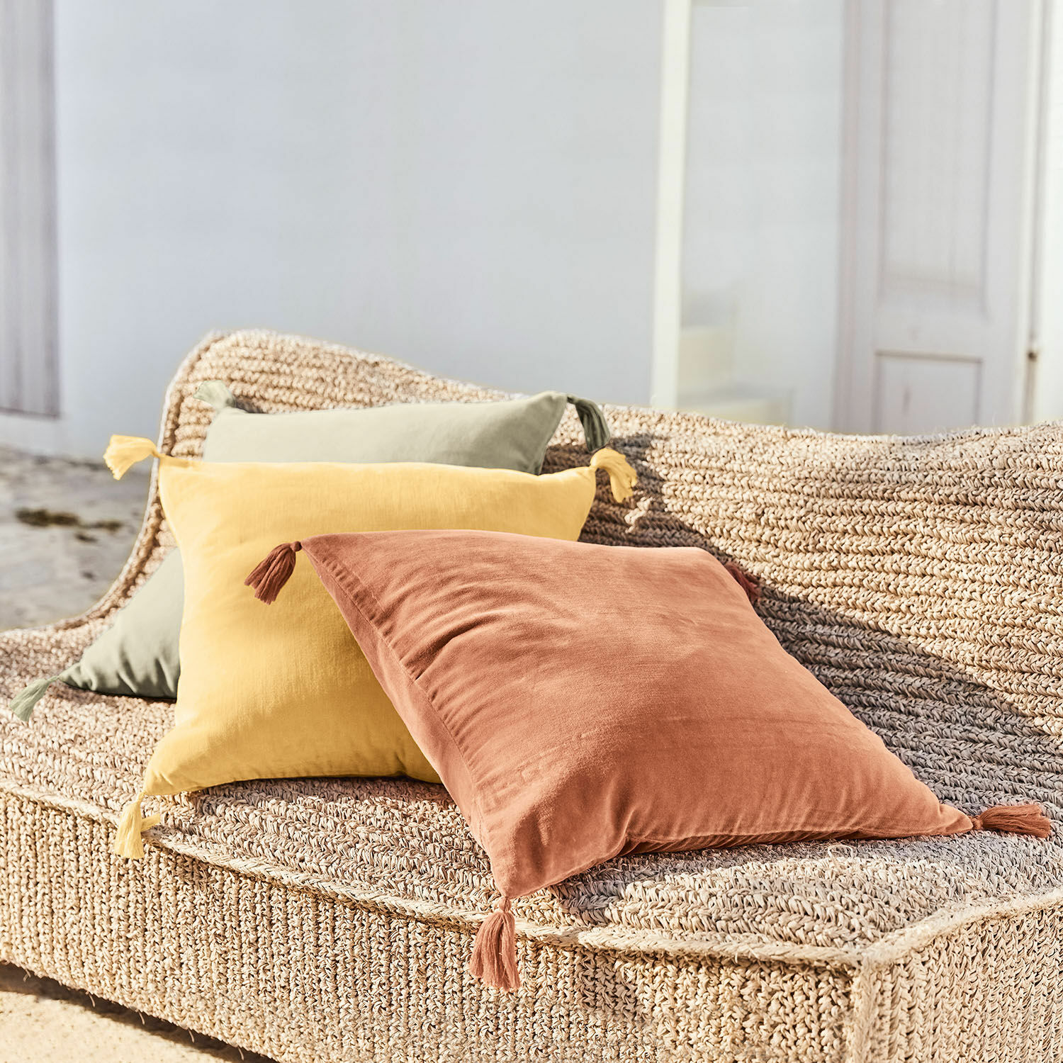 Drie decoratieve katoenen kussens in terracotta, geel en groen op een gevlochten zitmeubel buiten
