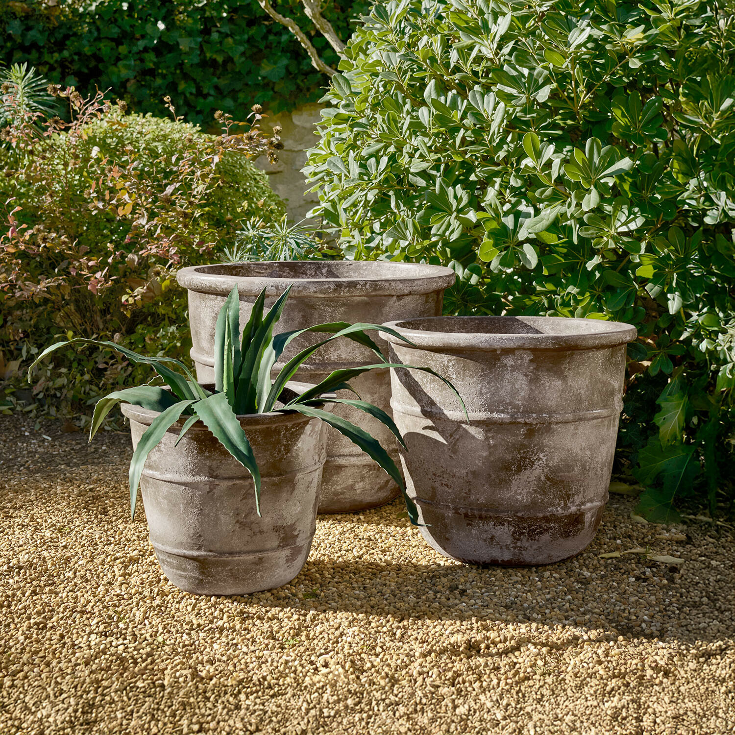 Lot de 3 cache-pots Arlivonne Trois grands pots en pierre grise sur un sol gravillonné dans un jardin avec des plantes vertes