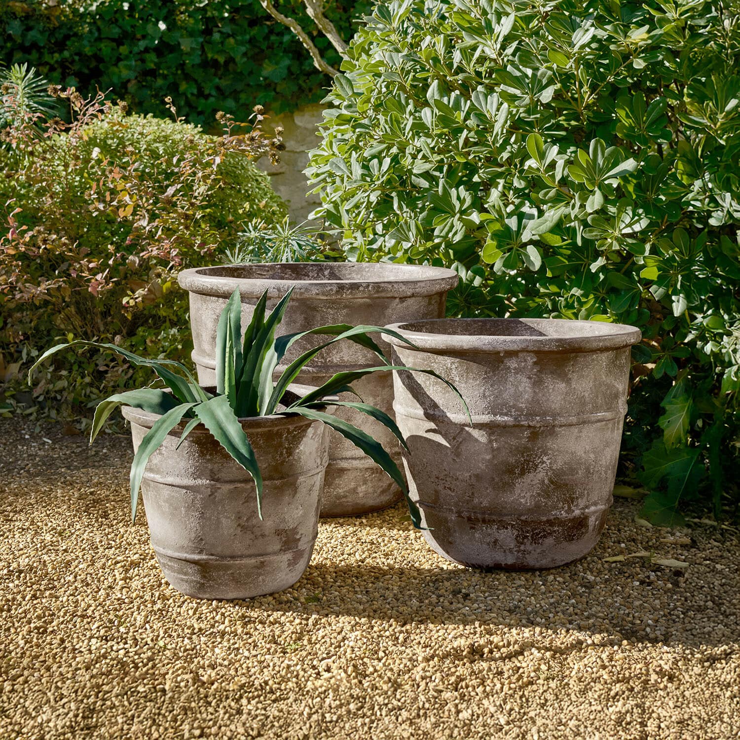 Lot de 3 cache-pots Arlivonne Trois grands pots en pierre grise sur un sol gravillonné dans un jardin avec des plantes vertes