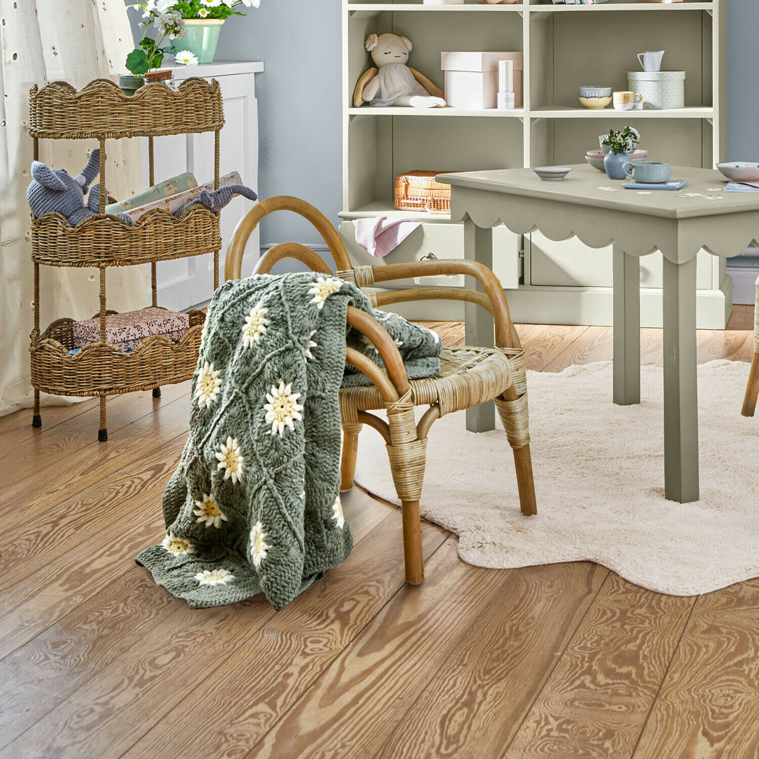 Chaise enfant Tinytreasure Plaid tricoté gris avec fleurs blanches sur chaise en osier dans une chambre d'enfant lumineuse au sol en bois