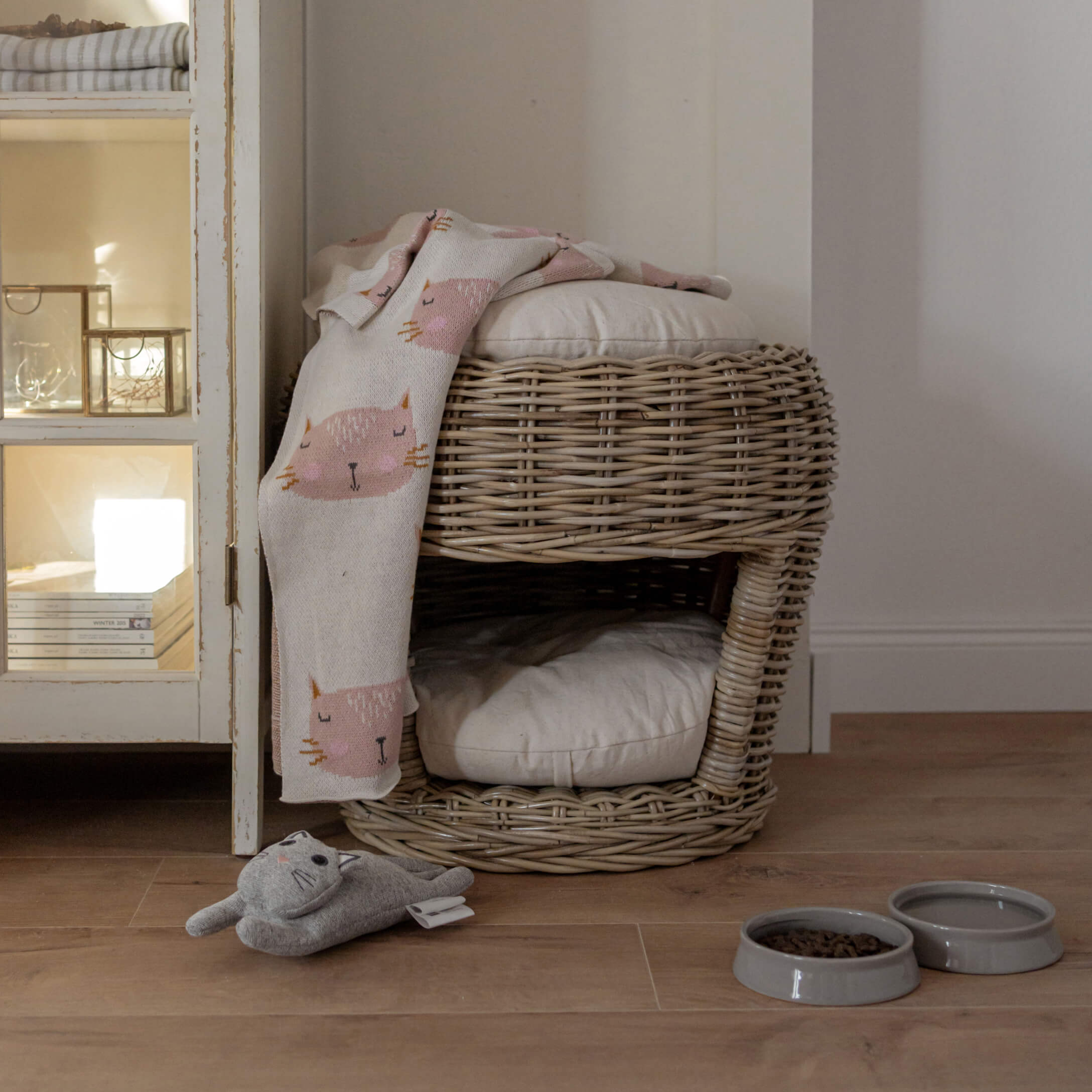 Niche double en rotin avec coussins clairs, couverture à motifs chats roses, jouet et gamelles pour chat