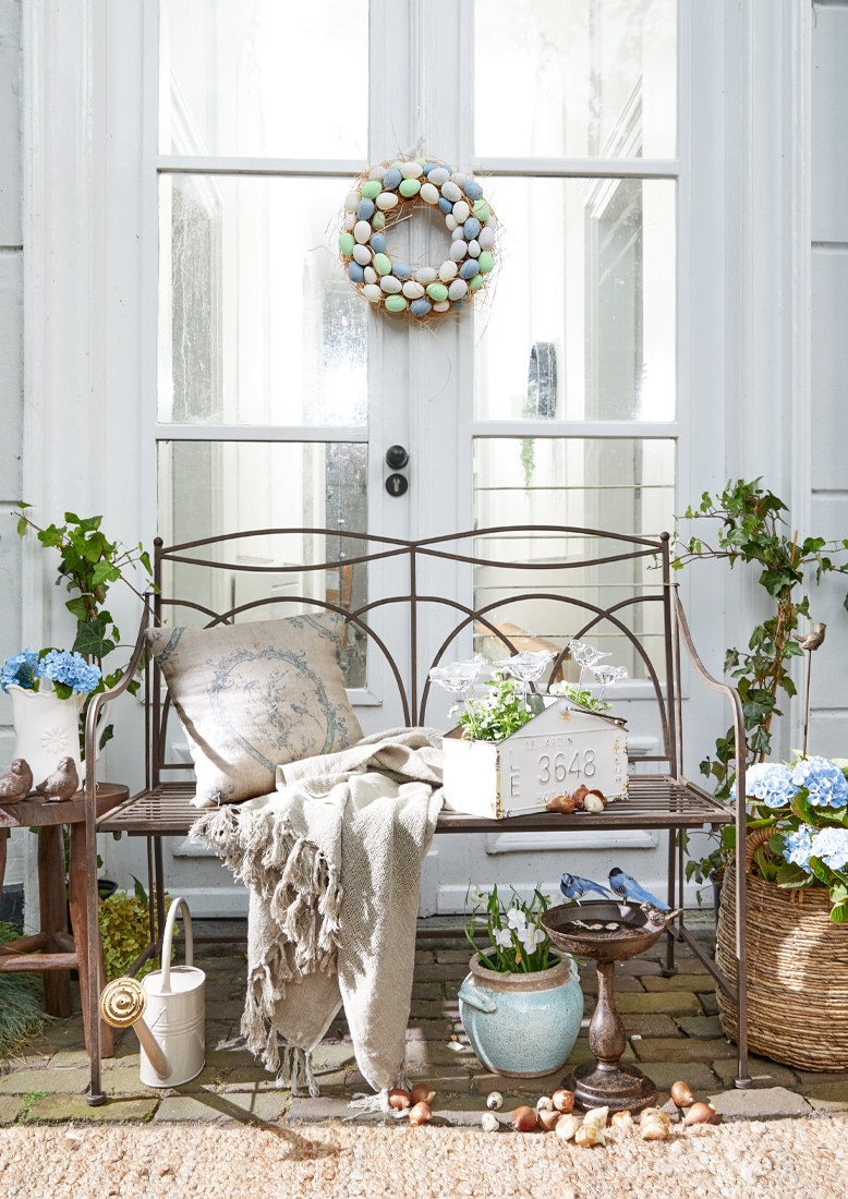 Metalen tuinbank met beige deken en kussen, bloemen en krans van pastelkleurige eieren