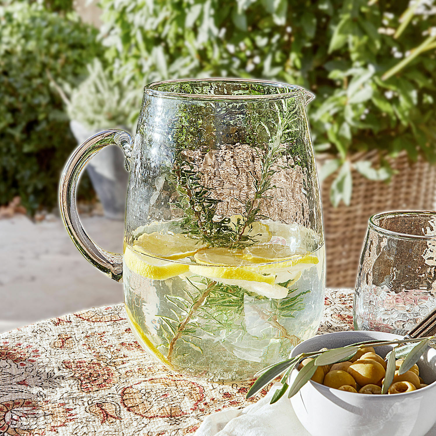 Glazen kan met water, citroen en kruiden op decoratief tafelkleed, zomerse drank op terras