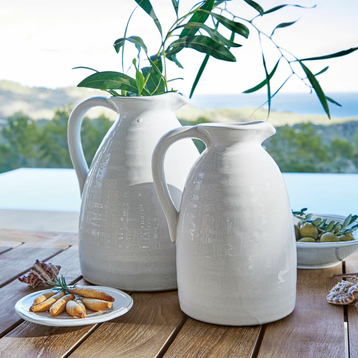 Lot de 2 vases Yarick Carafes blanches en céramique sur table en bois avec feuilles vertes et amuse-bouches, décoration