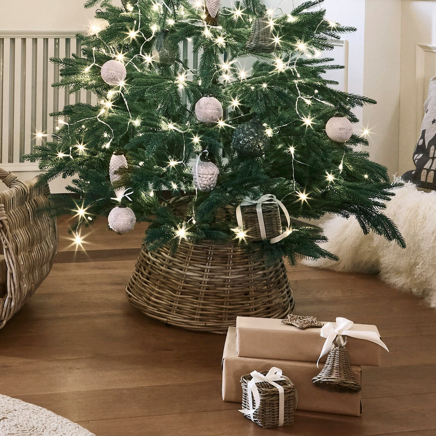 Tour de sapin de Noël Kiente Sapin de Noël dans un support en osier avec décorations naturelles et cadeaux emballés en papier brun sur un sol en bois