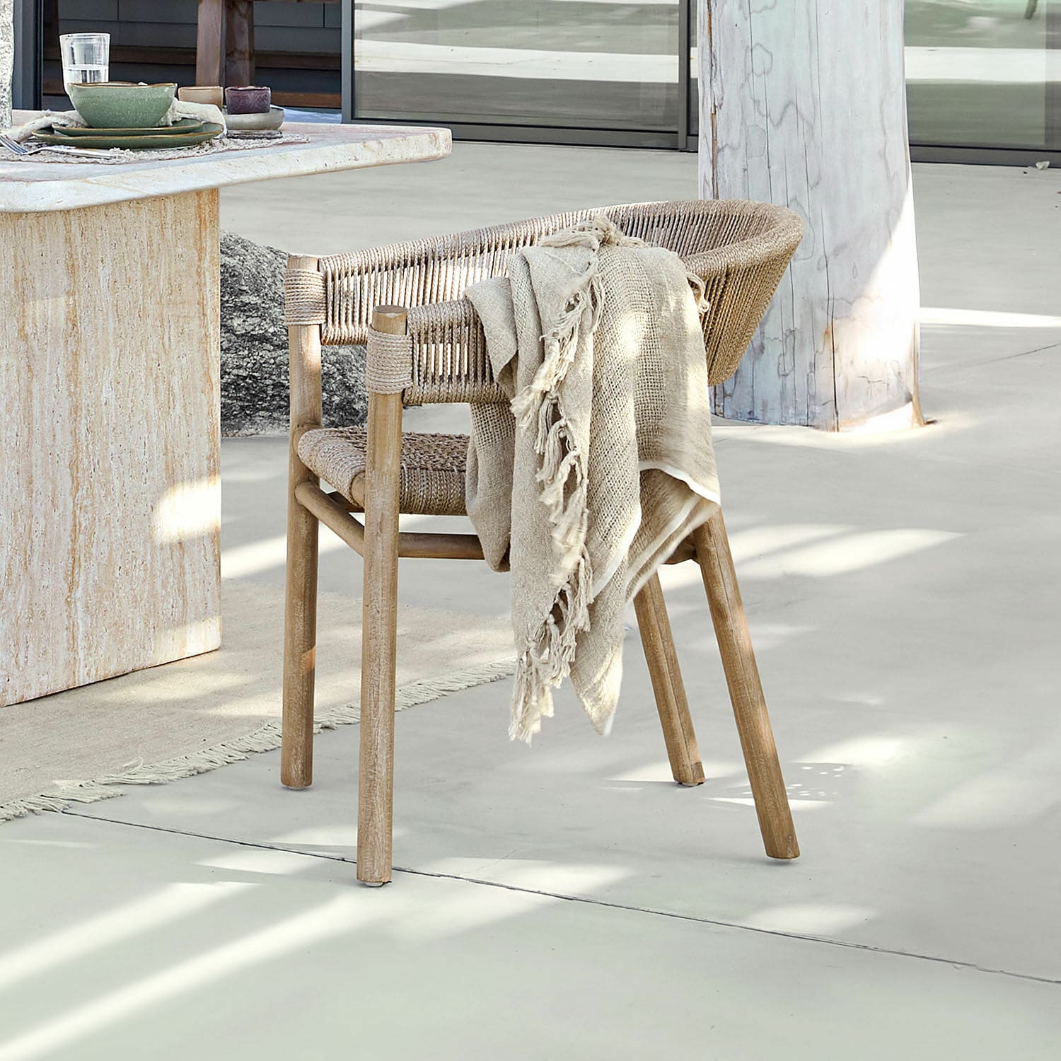 Fauteuil FSC®-100% Garmontique Chaise en bois et corde naturelle avec plaid beige sur sol clair près d'une table en pierre
