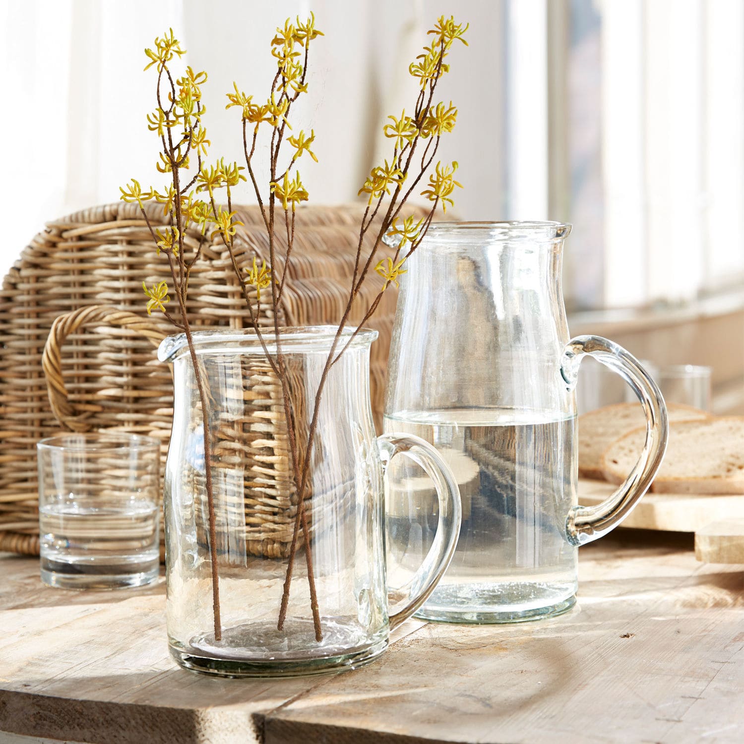 Carafe en verre transparent et pichet avec branches à fleurs jaunes sur table en bois clair