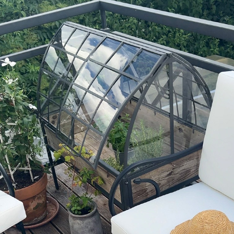 Kleine tuinkas met metalen frame en houten basis op terras met planten en tuinmeubilair