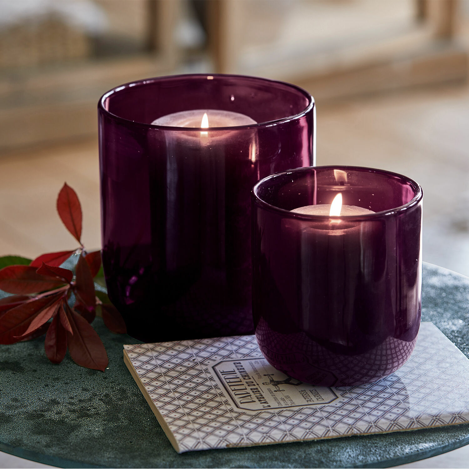 Windlicht set van 2 Theodor Twee paarse glazen windlichten met brandende kaarsen op een decoratieve onderzetter op een groene tafel