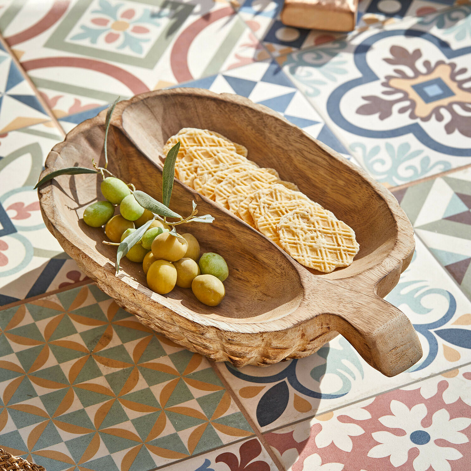 Bol en bois avec poignée contenant des olives vertes et des crackers sur un carrelage coloré