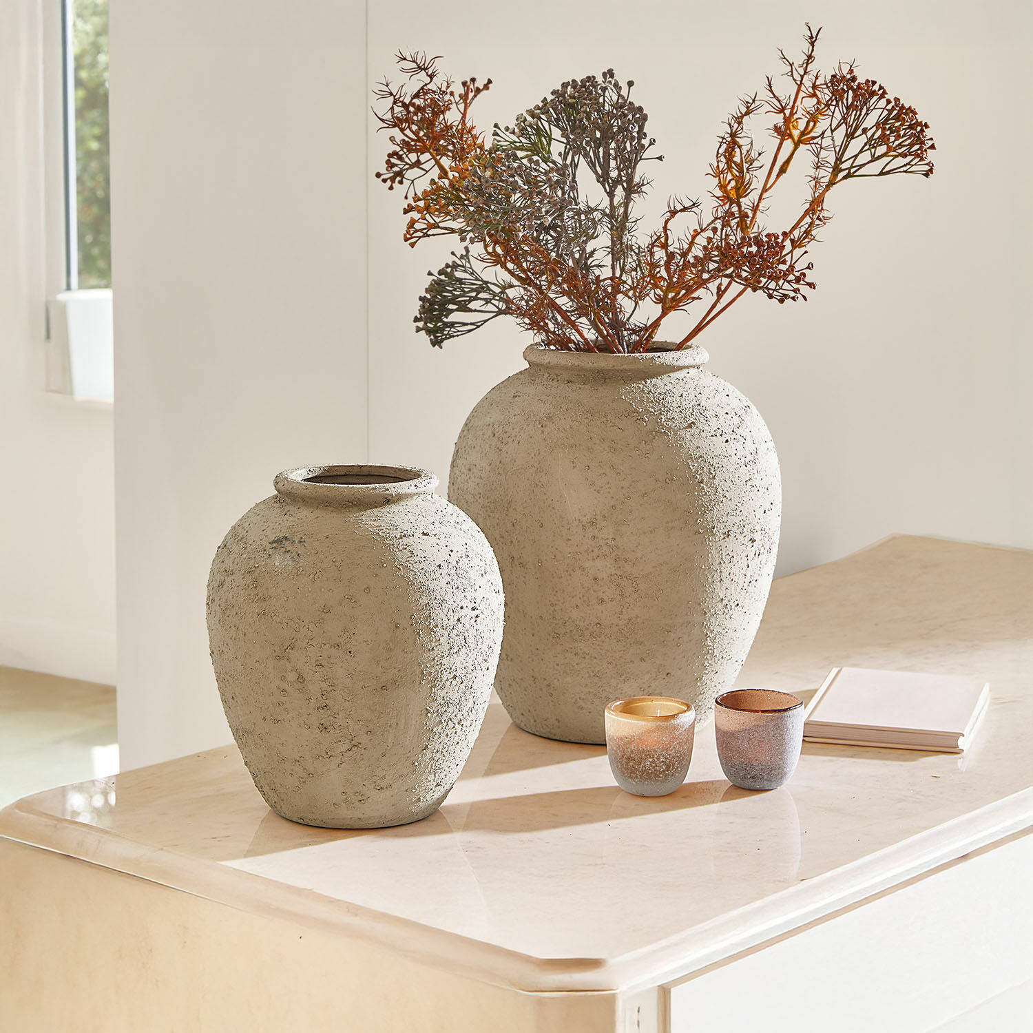Deux grands vases en béton gris sur un meuble clair avec branches sèches et bougies décoratives