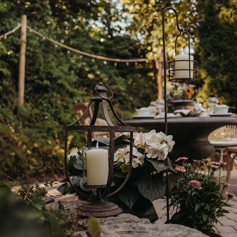 Metalen lantaarn met witte kaars op stenen muurtje in tuin met bloemen en tafel op de achtergrond
