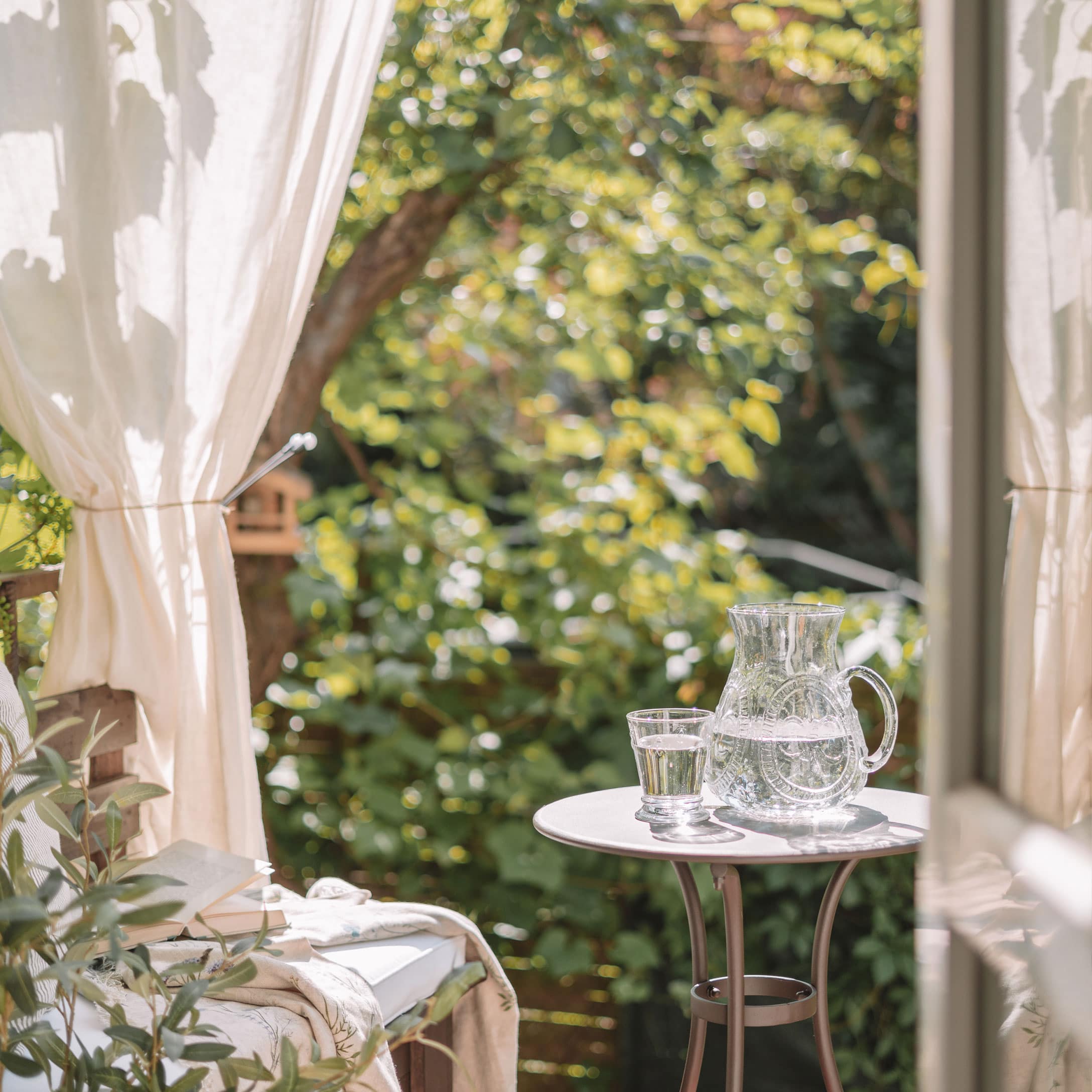 Glazen karaf en glas met water op witte tafel op terras met groene planten op de achtergrond