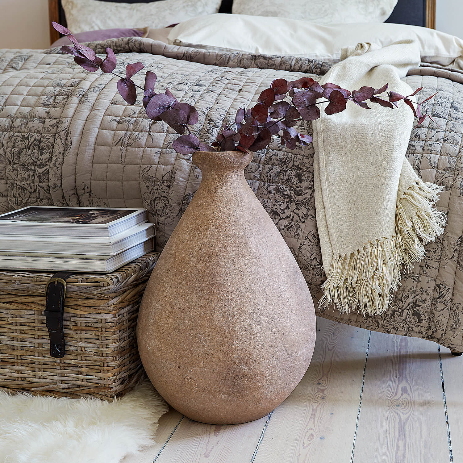 Grand vase en céramique beige avec branches séchées devant un lit et un panier en osier