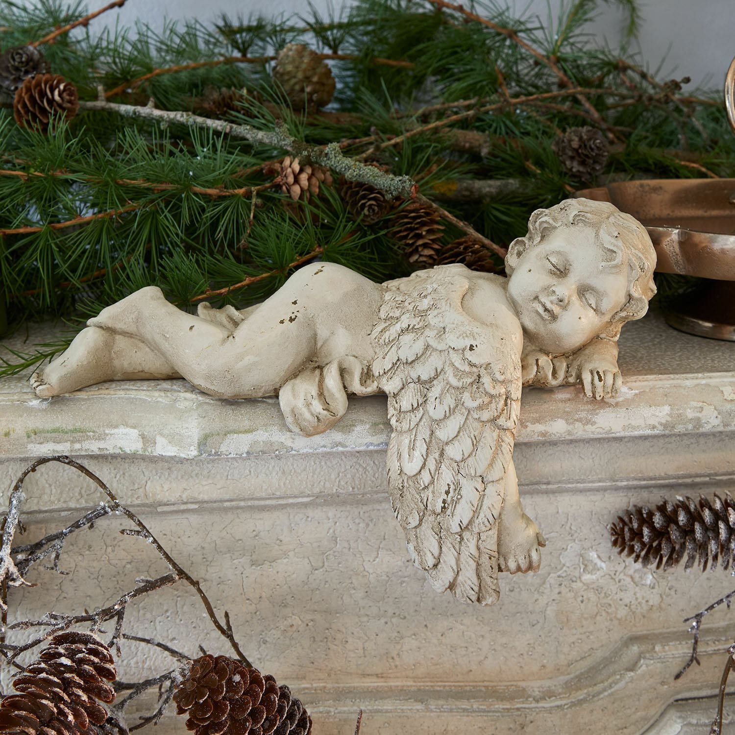 Figurine d'ange en béton crème posée sur une cheminée avec branches de sapin et pommes de pin, décoration