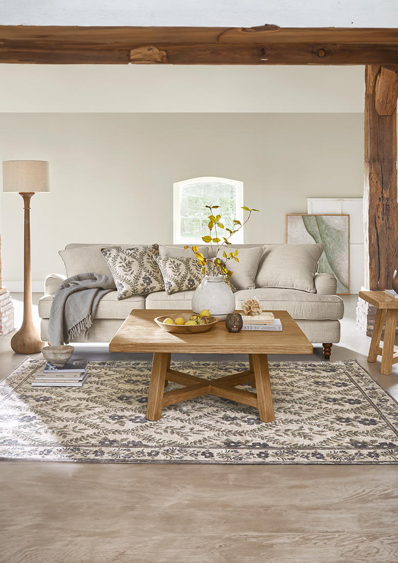 Beige stoffen bank met decoratieve kussens en houten salontafel op patroon vloerkleed in lichte woonkamer