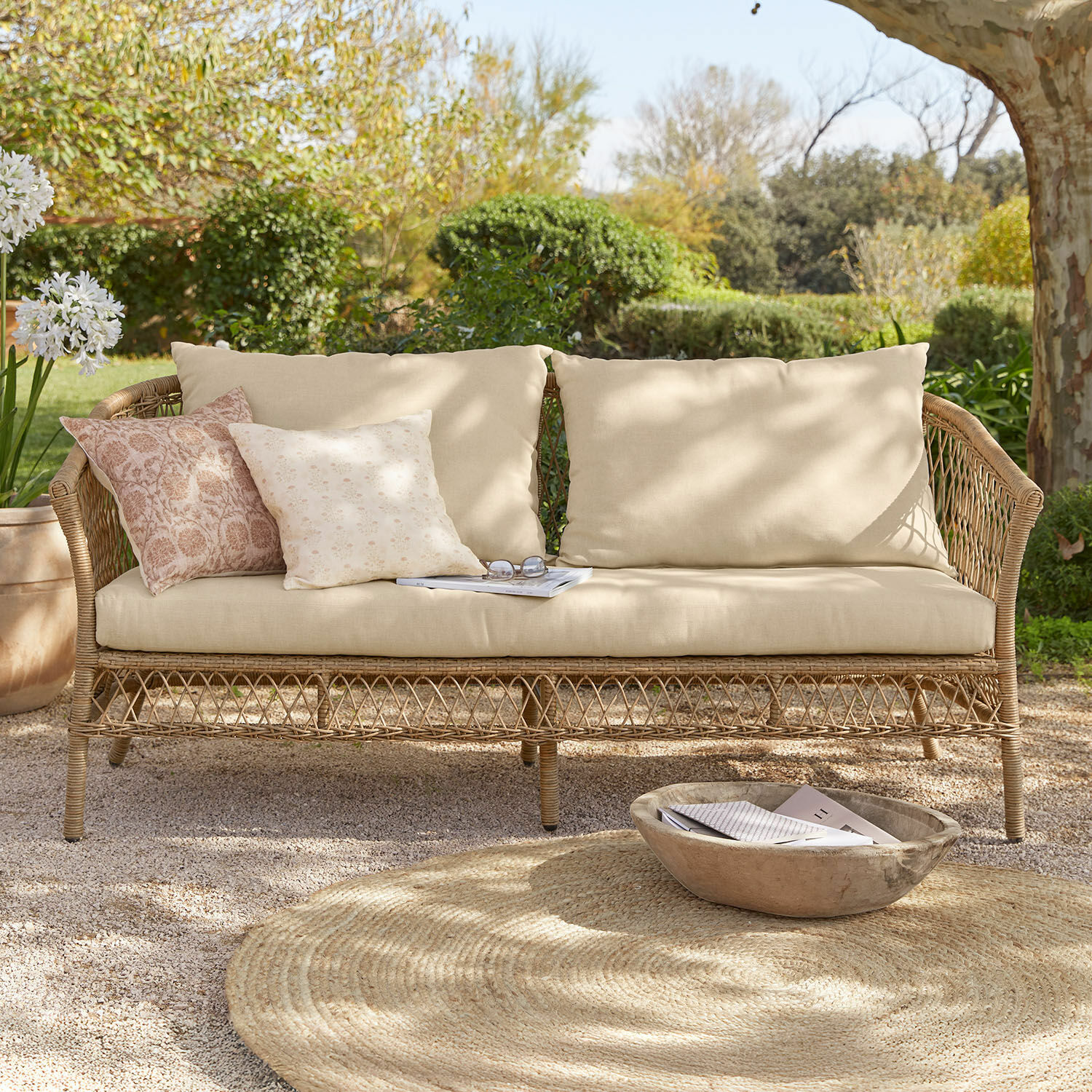 Canapé en rotin avec coussins beige et coussins décoratifs pastel dans un jardin ensoleillé