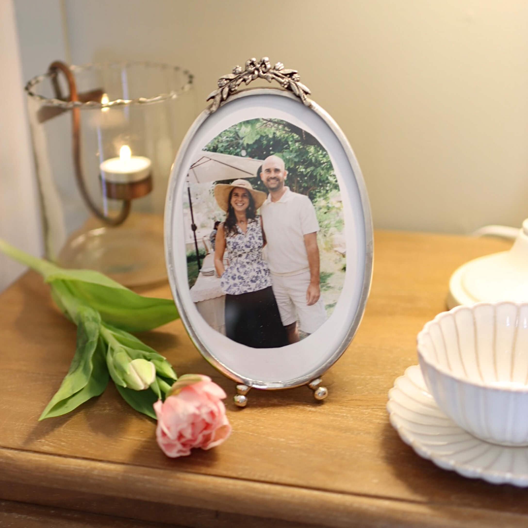 Cadre photo ovale en argent avec ornement floral sur une table en bois avec une rose et un photophore
