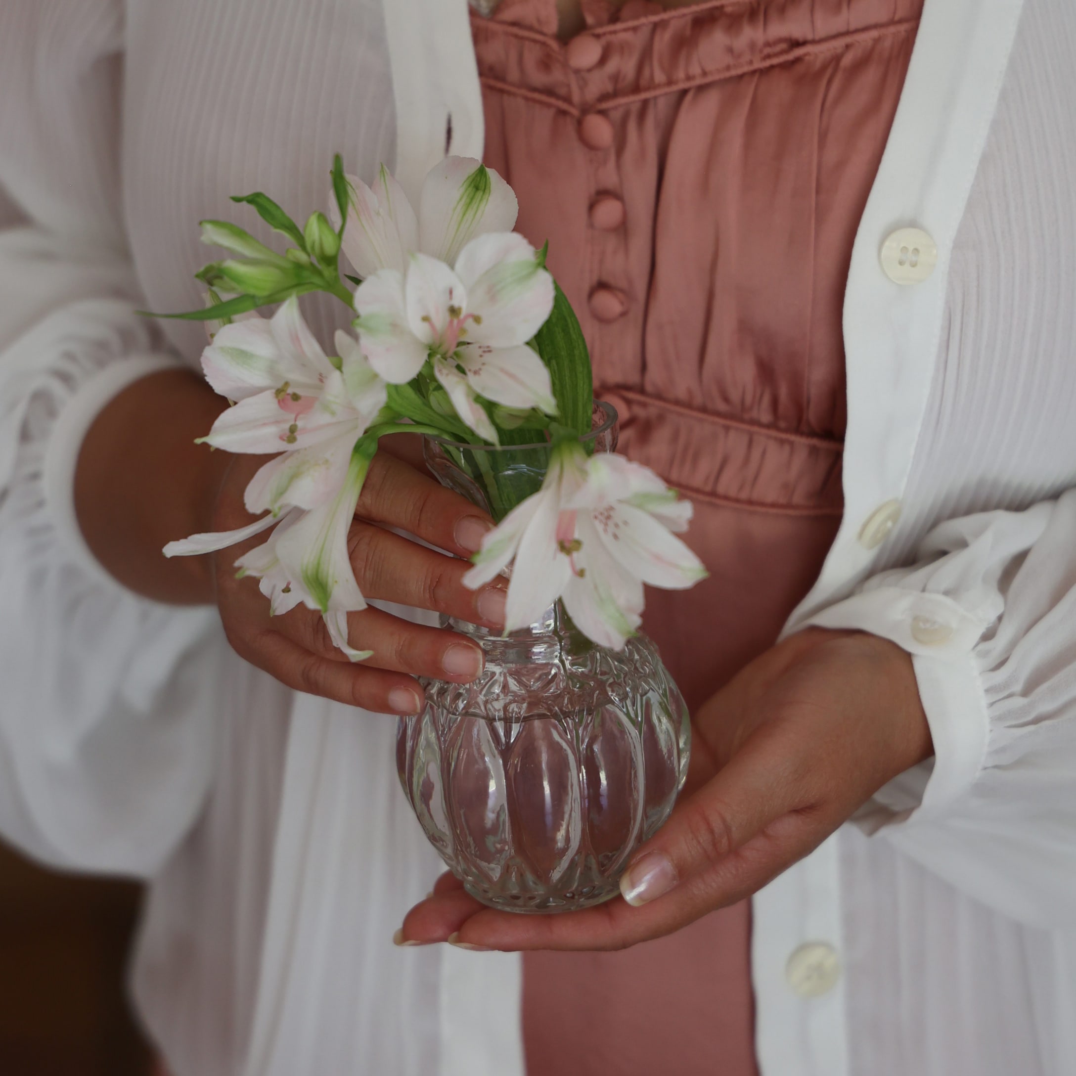 Vrouw houdt een doorzichtige glazen vaas met witte bloemen voor een roze jurk en witte cardigan