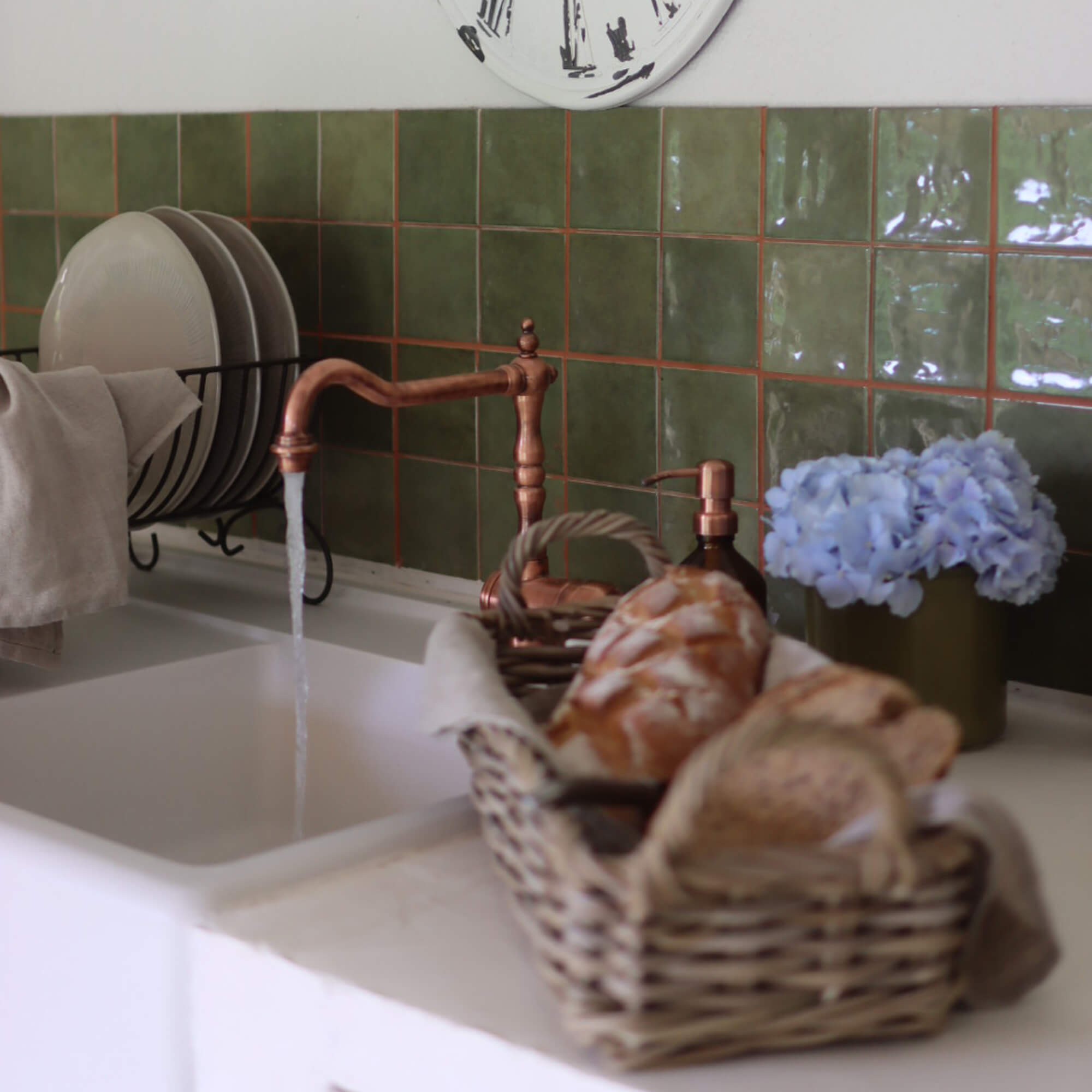 Cuisine avec robinet en cuivre, carreaux verts, panier de pain et fleurs bleues décoratives