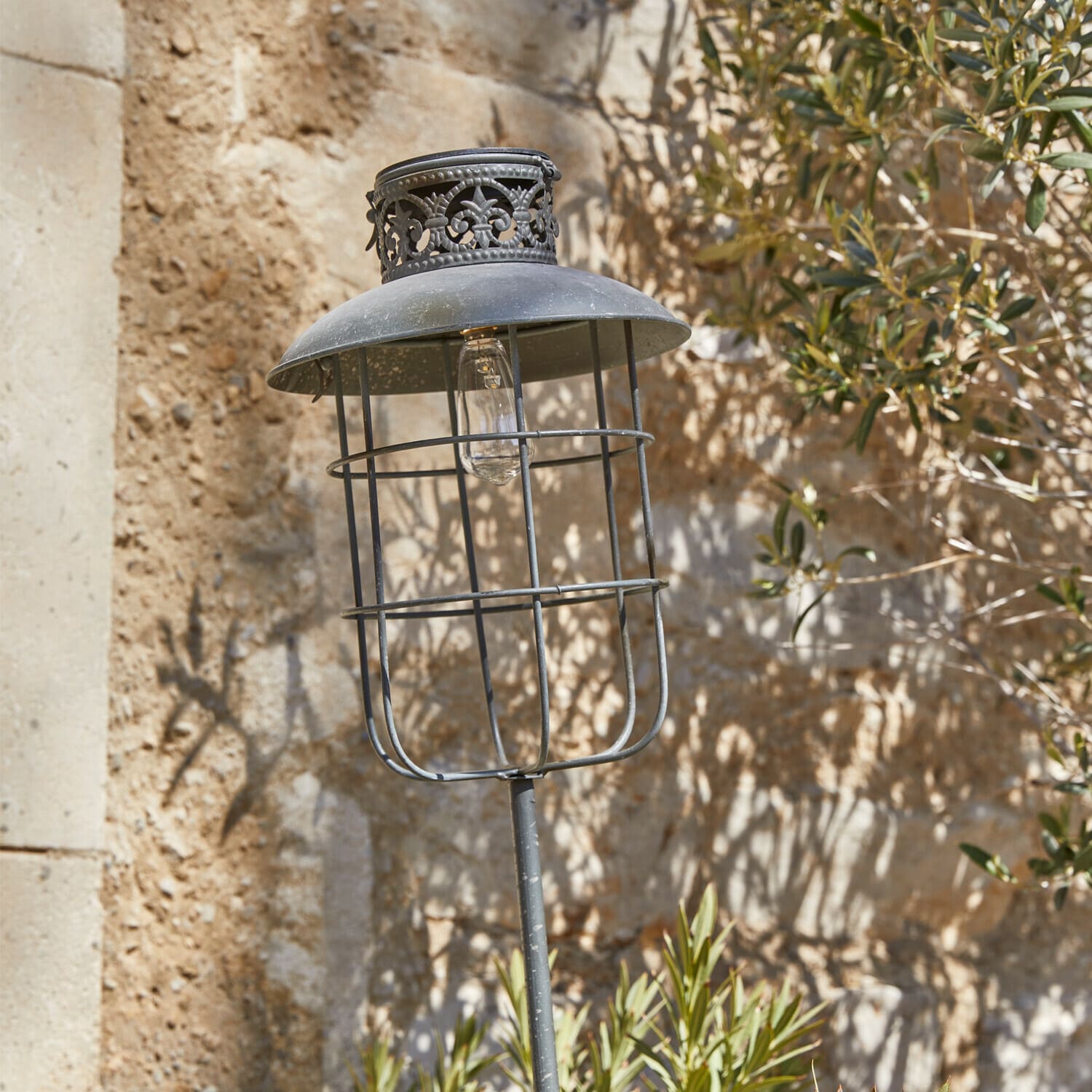 Lanterne solaire Nistos Lampe de jardin en métal noir avec abat-jour décoratif et ampoule visible sur fond de mur en pierre