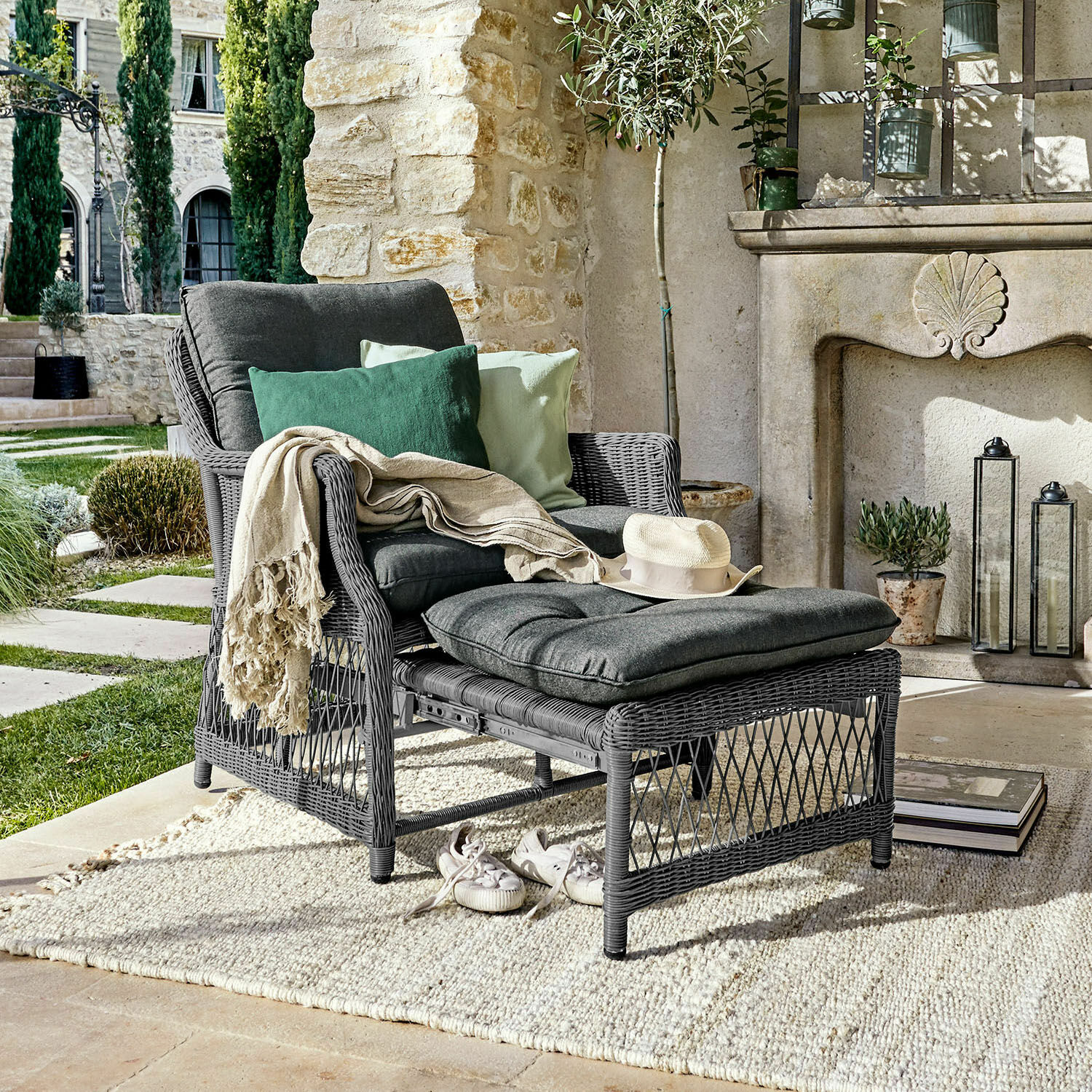 Fauteuil Howe Grijze rotan fauteuil met voetenbank en donkergrijze kussens, versierd met groene kussens op een terras