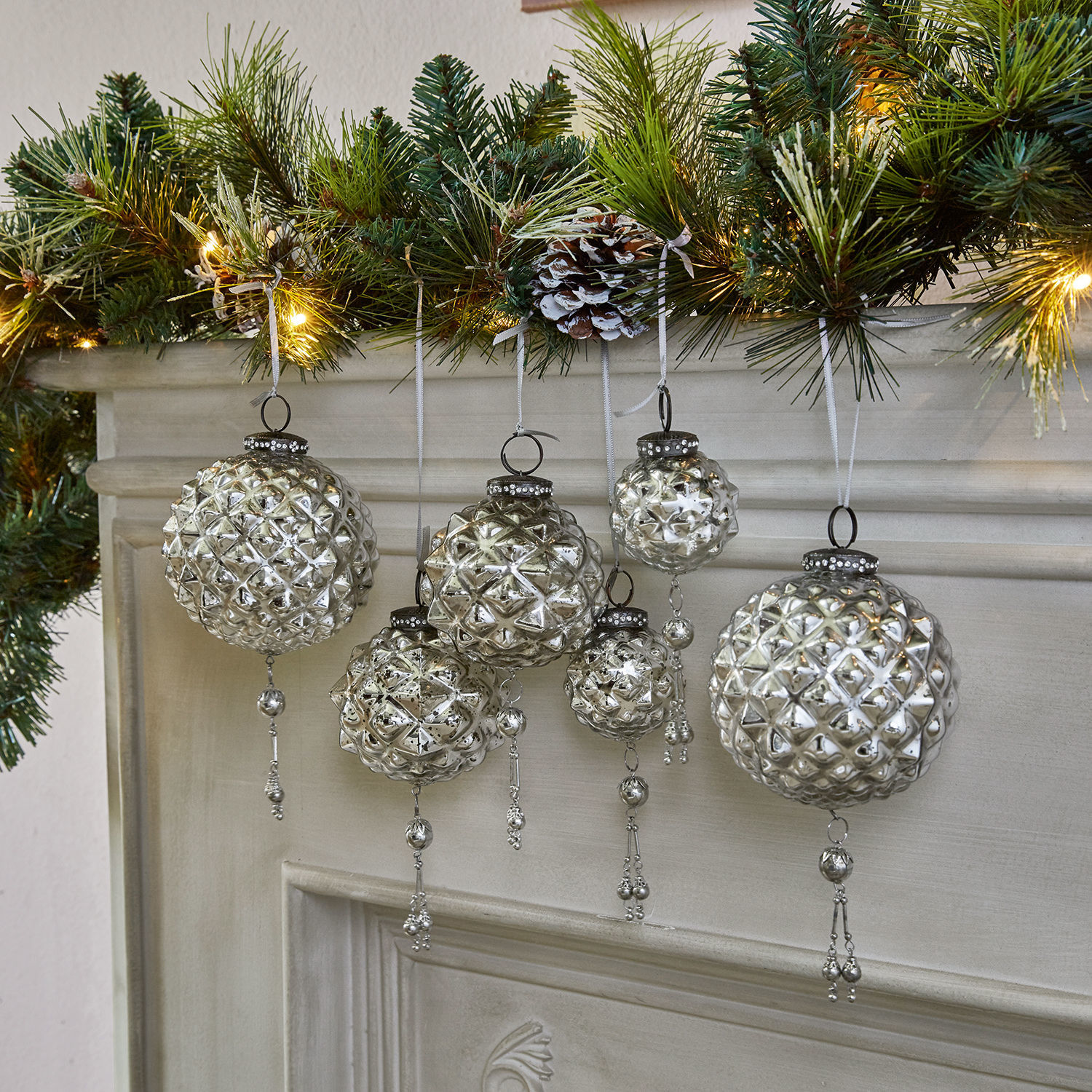 Boules de Noël en verre argenté avec décorations suspendues devant une cheminée et des branches vertes éclairées