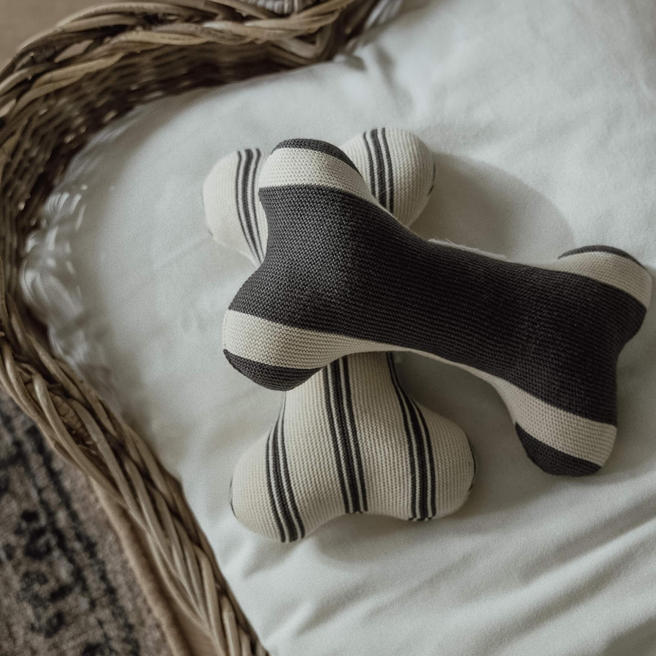 Deux jouets en forme d'os en tricot noir et blanc rayé sur fond blanc dans un panier en osier