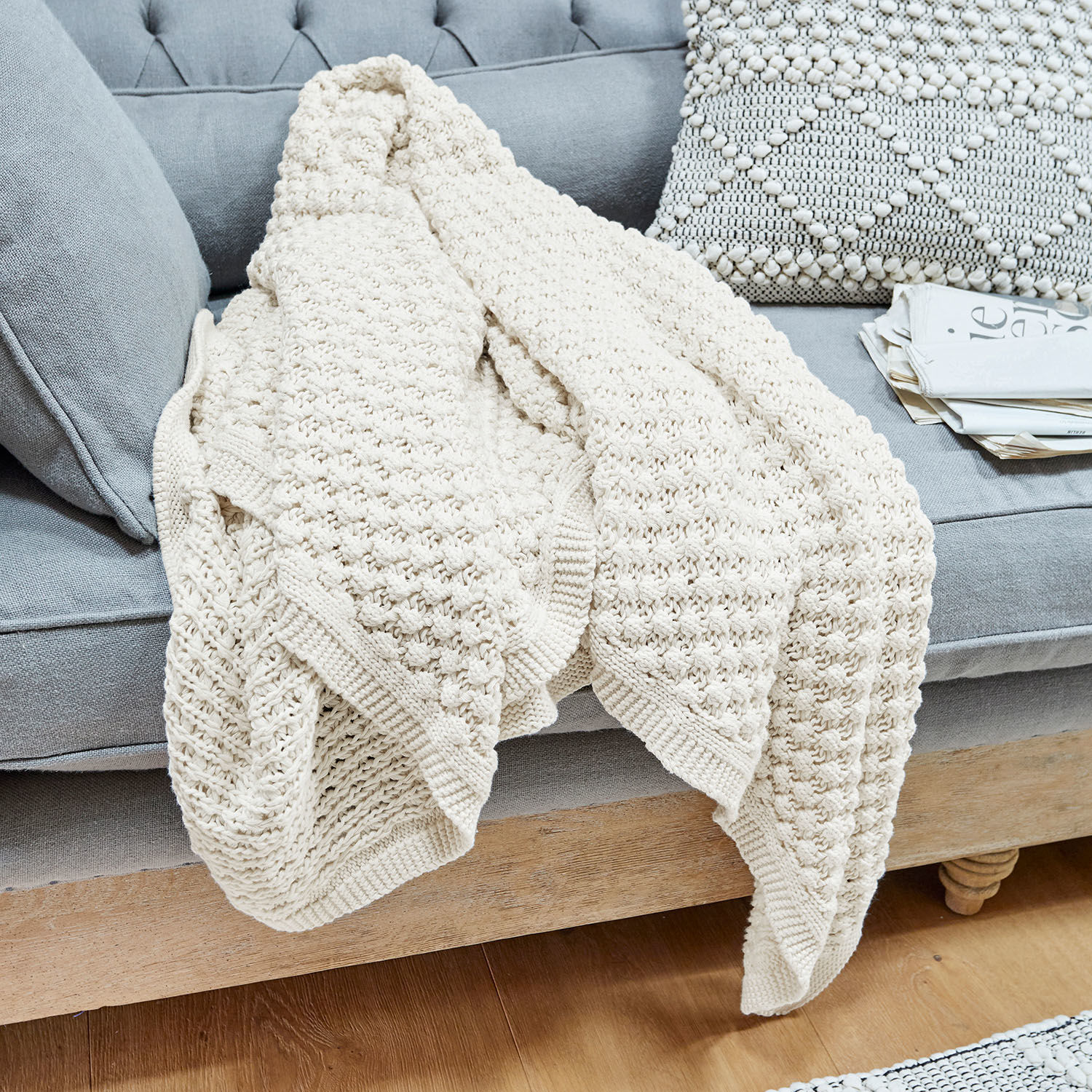 Plaid Tremes Plaid crème en coton tricoté posé sur un canapé gris avec coussin décoratif et journaux