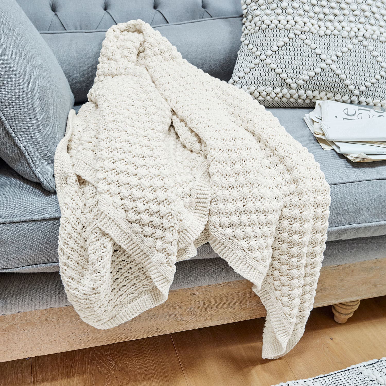 Plaid Tremes Plaid crème en coton tricoté posé sur un canapé gris avec coussin décoratif et journaux