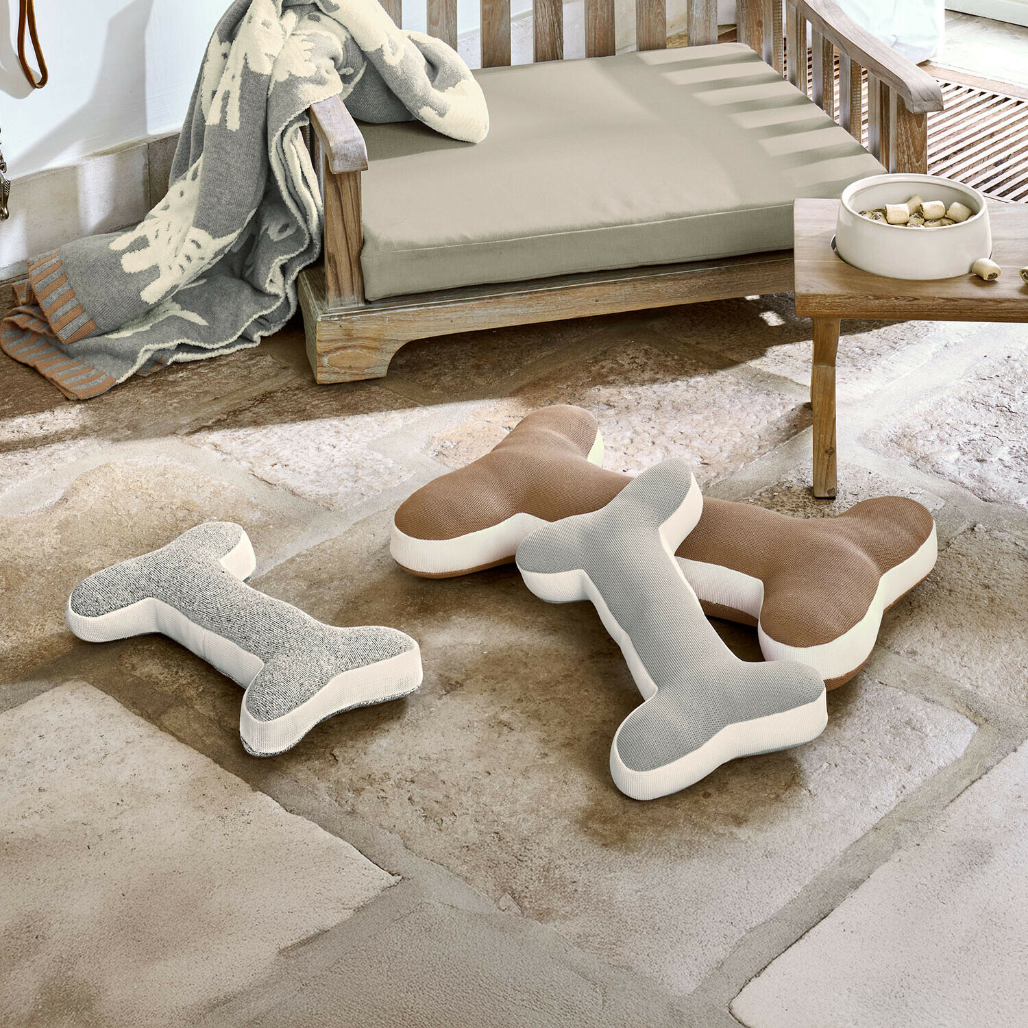 Lot de 3 coussins Fluffido Trois coussins en forme d'os gris et marron sur un sol en pierre près d'un banc en bois avec coussin beige