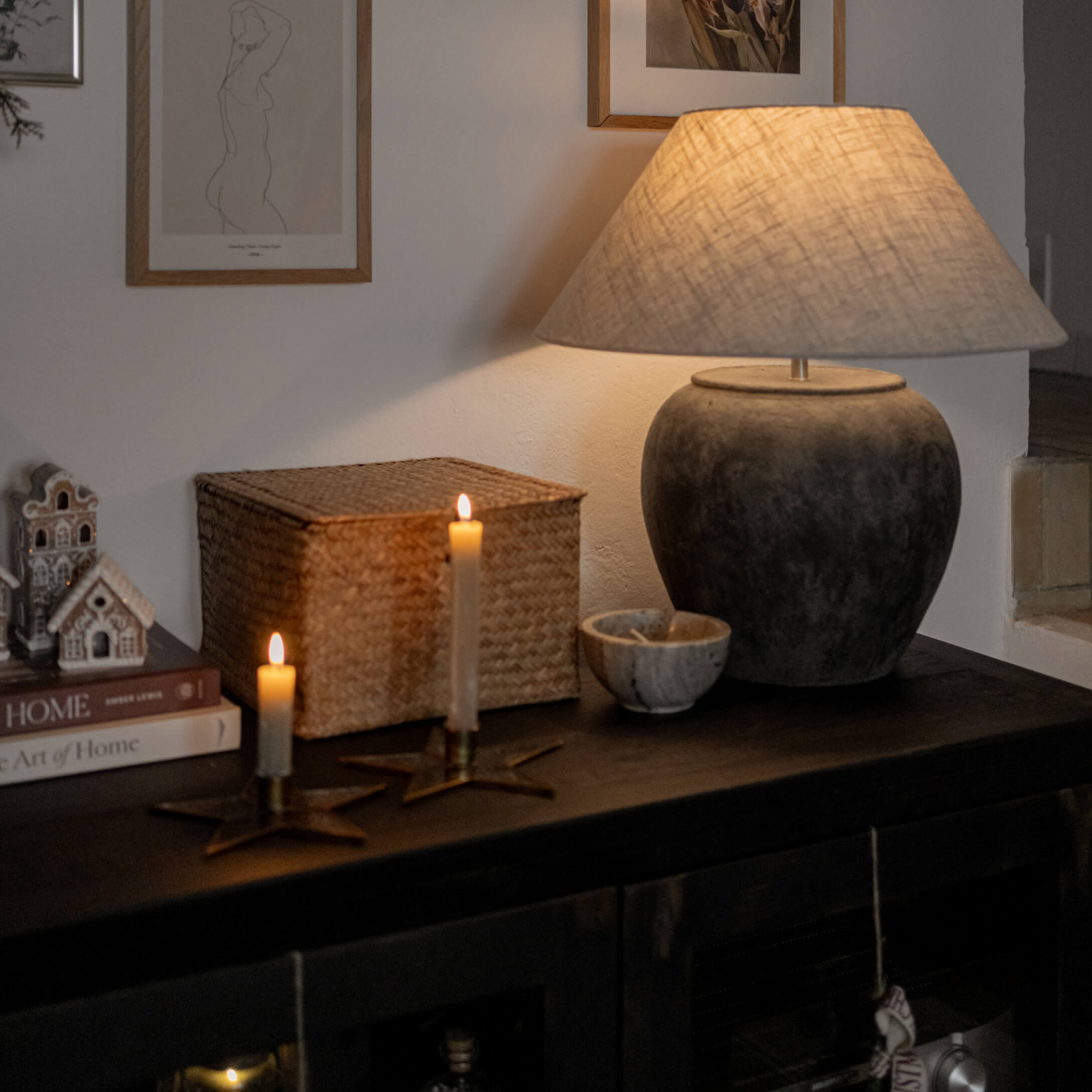 Tafellamp van donkergrijs keramiek met beige kap op donkere kast met kaarsen en decoraties