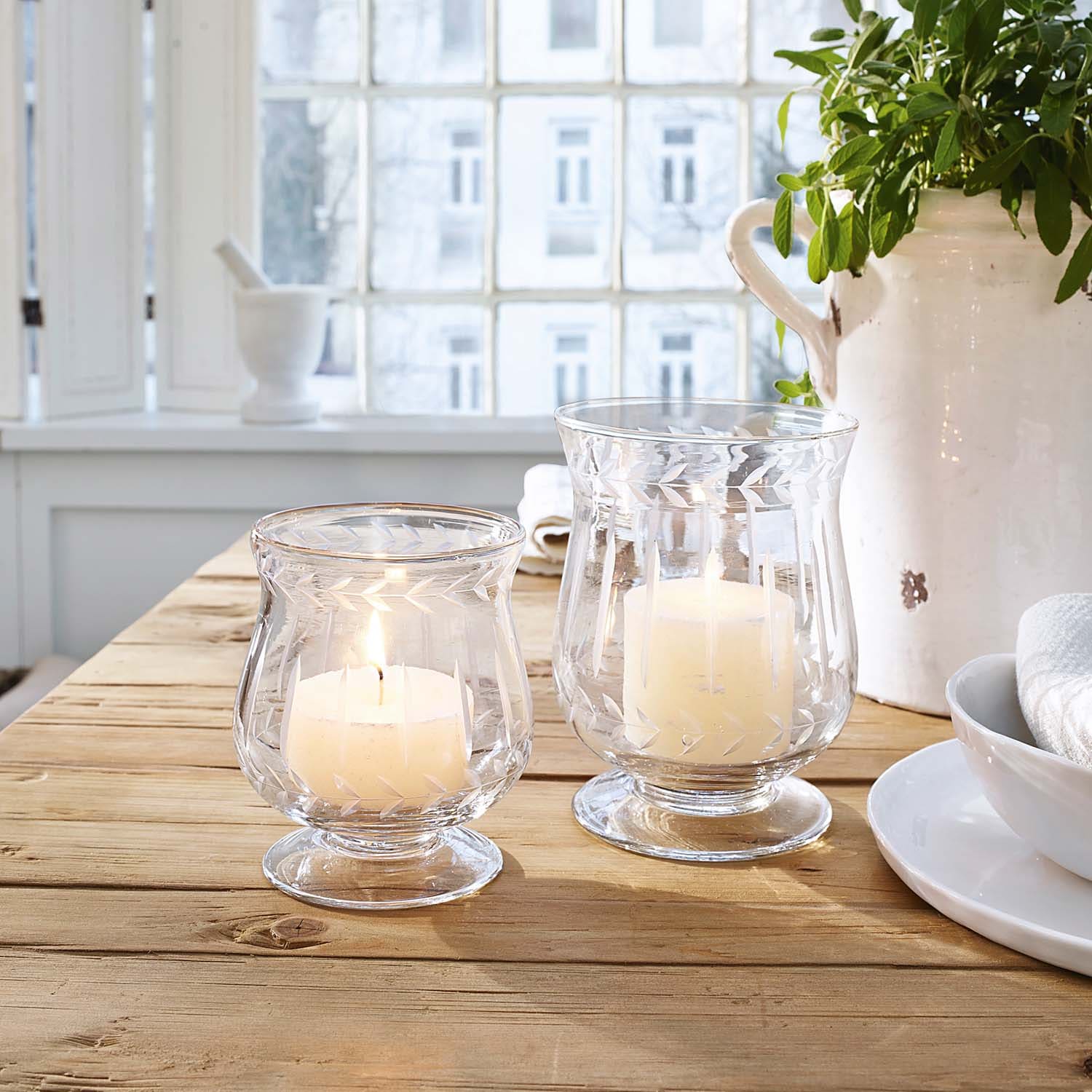 Twee glazen windlichten met witte kaarsen op houten tafel in lichte kamer