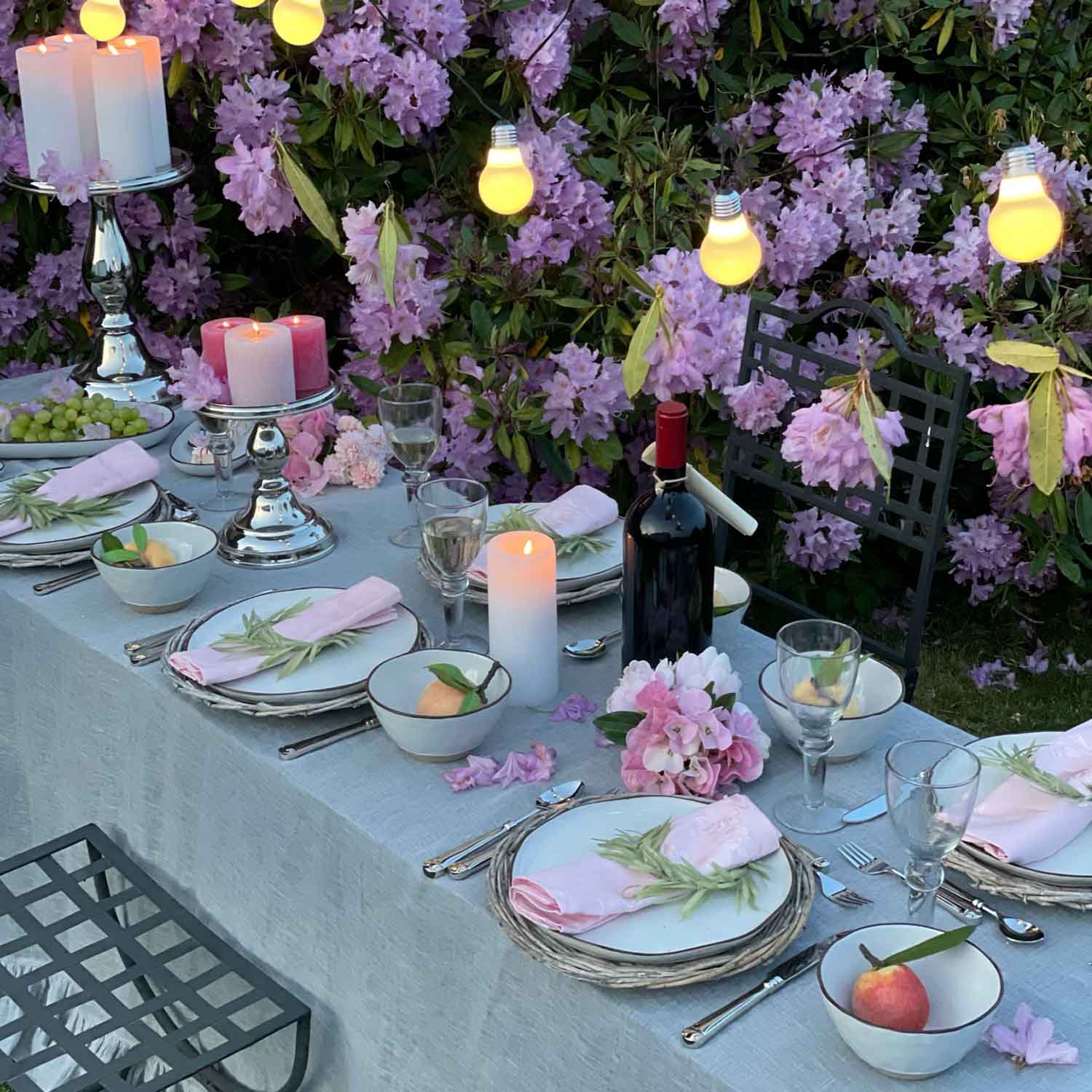 Table de jardin élégamment dressée avec serviettes roses, bougies et bouteille de vin devant des fleurs violettes