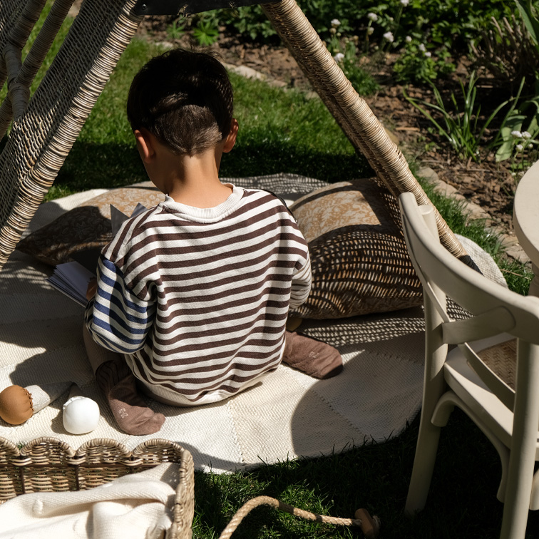 Enfant en pull rayé assis sur un tapis sous une tente en osier dans un jardin, moment de détente